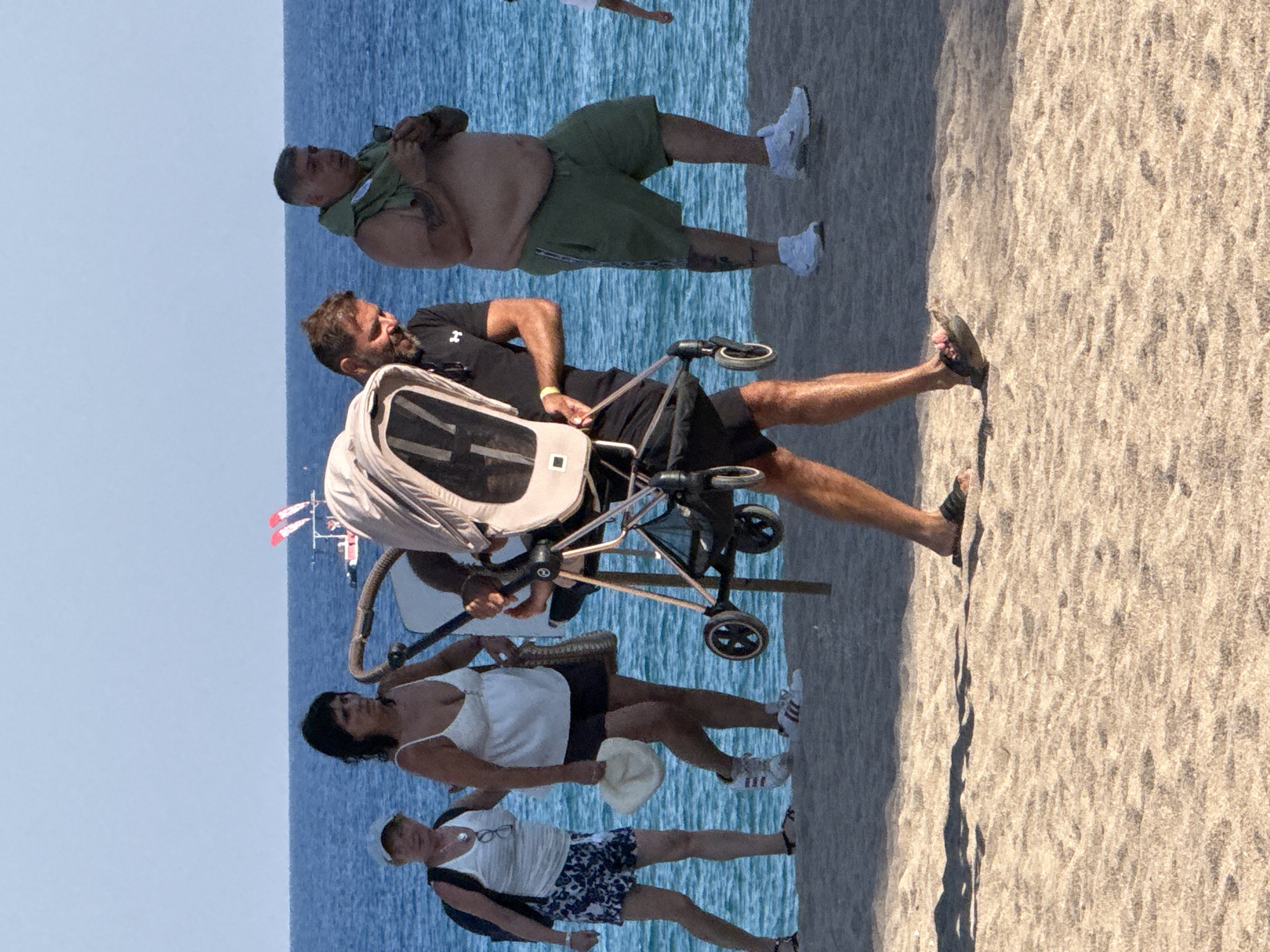 The image captures a sunny day at a beach in Blanes, Spain, with the ocean in the background and sandy terrain in the foreground. Several people are present, engaged in various activities.
In the upper portion of the frame, a man in green shorts and a light brown hoodie stands near the water, holding his clothing. Below him, another man with a beard and a black t-shirt is seen with a stroller. The stroller is open and appears to be pushed by a person whose arm is visible. Further down, a woman with dark hair wears a white top and dark shorts, holding a light-colored hat. Behind her, another woman in a white top and patterned shorts, wearing glasses, is also visible.
The scene suggests a casual outing, possibly a family or friends enjoying a day at the beach. The bright sunlight indicates it's daytime, and the clear blue of the ocean hints at fair weather. The presence of a stroller and beach attire suggests a relaxed atmosphere.
There is no visible text in the image. The overall impression is one of a pleasant summer day at the Spanish coast.