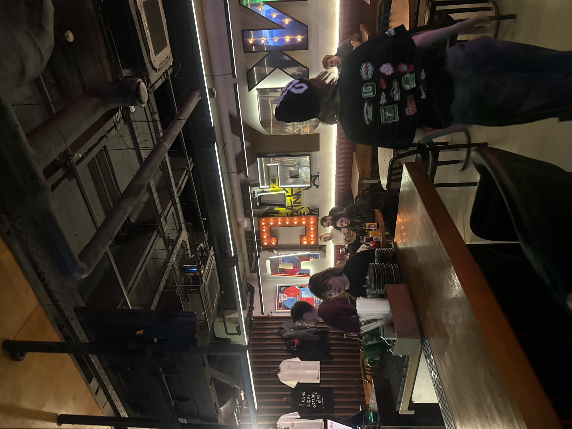 The interior of a Moscow restaurant in Russia shows patrons seated at tables along a counter, with a person wearing a black hat and jacket walking past. Neon letter signs and framed pictures decorate the walls, and various T-shirts hang for sale near the service area. The ceiling features exposed industrial piping and equipment.