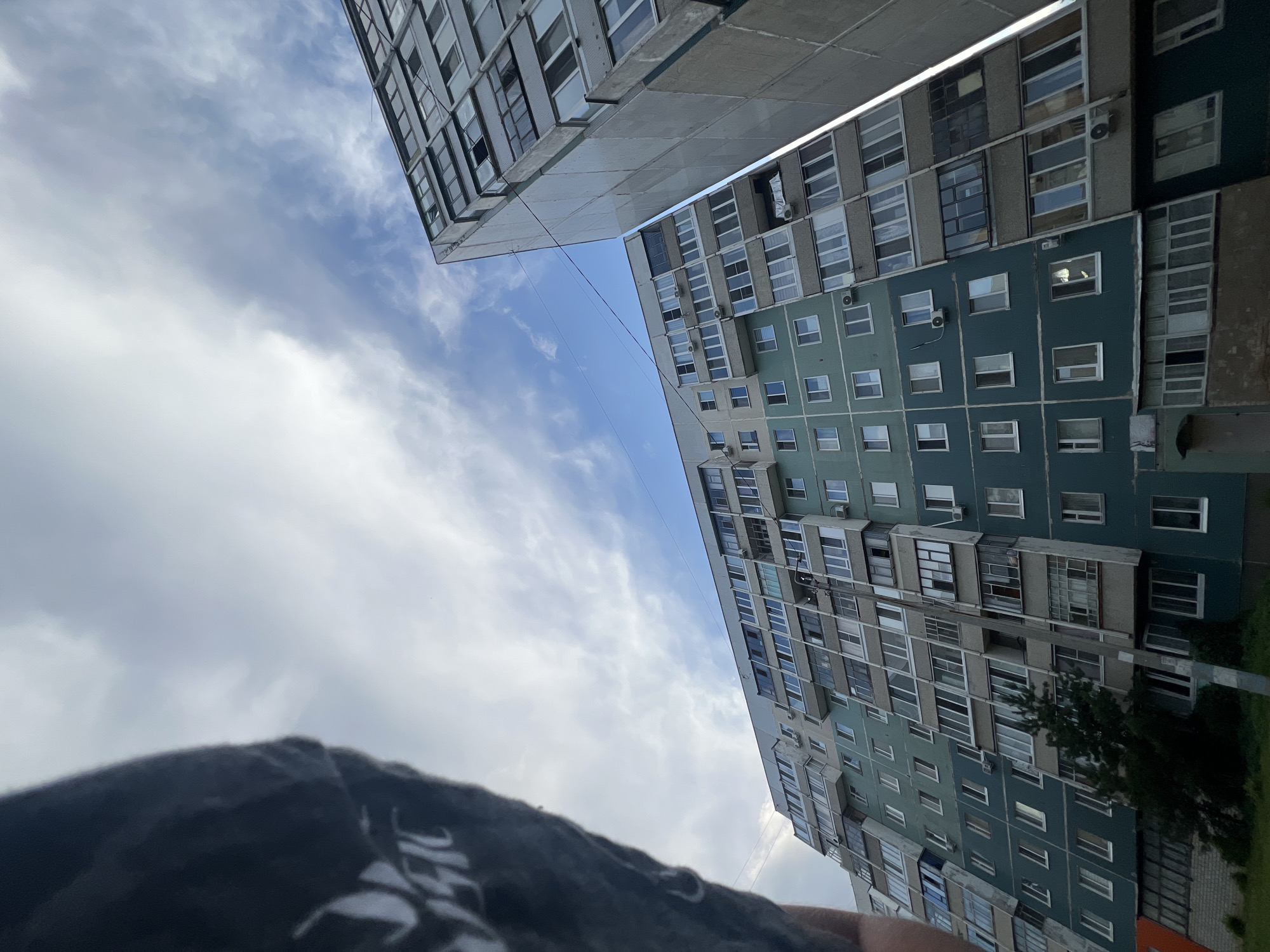 The image shows two apartment buildings in Ulyanovsk, Russia, viewed from a low angle looking up at the sky. The building in the foreground is a multi-story apartment complex with a predominantly teal facade, featuring numerous balconies and windows. The building to the right is taller and appears to be constructed of concrete with a more industrial aesthetic. The sky is cloudy with patches of blue visible. The left edge of the image is partially obscured by a dark garment, possibly a sleeve or shoulder of a person wearing dark clothing with white lettering. The overall scene depicts an urban environment under a cloudy sky.