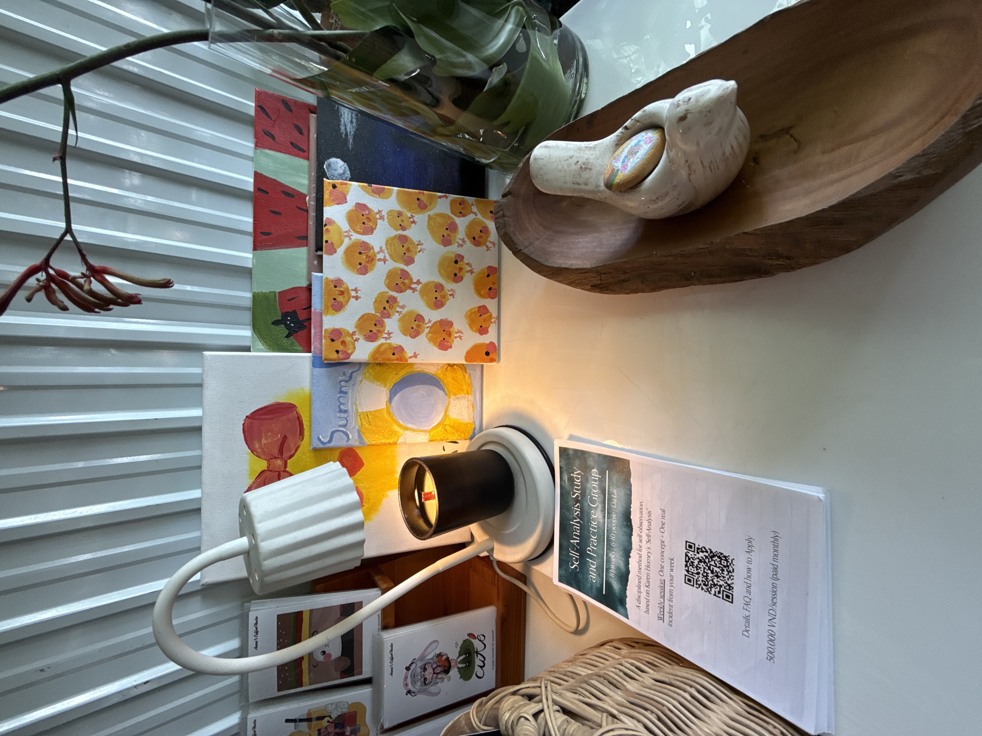 This image features a collection of various objects arranged together. A wooden dish containing a bird figurine sits near a stack of small canvases with paintings, including one with a chick pattern and another with a summer theme. A flexible arm lamp is positioned to illuminate the stack. Several business cards from "Anna's Coffee House" are visible. A brochure advertises a "Self-Analysis Study and Practice Group" in Da Lat, Vietnam, with details about the program and costs. The arrangement of the objects suggests a display or a temporary collection.