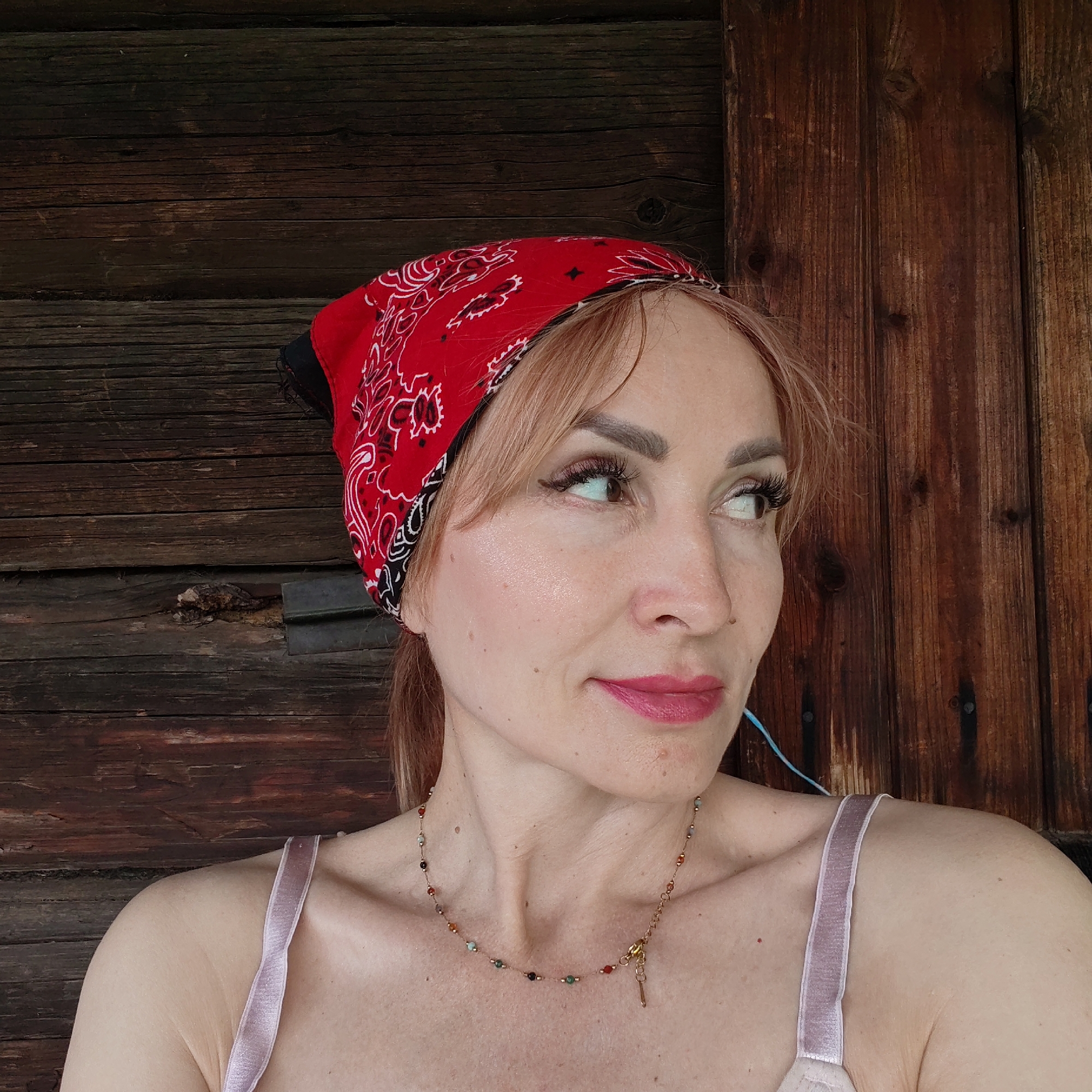 A woman with fair skin, reddish-blonde hair, and wearing a red paisley patterned bandana is pictured from the chest up. She is wearing a necklace and a light pink top with narrow straps. She is looking to her right. The background is a weathered wooden wall or door, likely an exterior. The image does not contain any information that could identify the location as Chaikovsky, Russia.