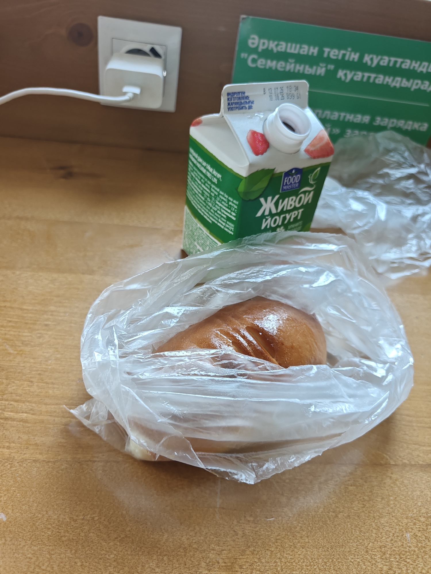 The image shows a close-up view of a wooden table with a white power adapter plugged into a white wall socket. To the right of the power adapter is a carton of "Живой йогурт" (Live Yogurt) with strawberries depicted on it, and a clear plastic bag containing a golden-brown baked good. In the background, a green sign with text in Kazakh and Russian is visible. The text on the green sign mentions "ркашан тегін қуаттандыр" which translates to "always free charging" and a brand name in quotes, possibly indicating a location in Petropavl, Kazakhstan that offers free charging services. The overall scene suggests a casual setting, possibly a cafe or a public space.