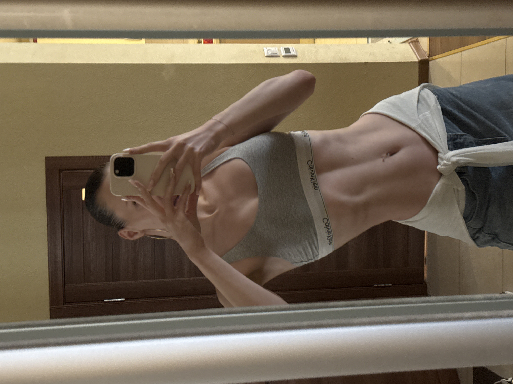 This media file depicts a person taking a mirror selfie in what appears to be a bathroom. The person is facing the mirror, holding a smartphone to capture the reflection. They are wearing a grey sports bra with "Calvin Klein" branding and blue jeans. A white garment is tied around their waist, over the jeans. The background shows a wooden door and textured walls, consistent with an indoor setting. There are light fixtures visible on the wall above the door. The lighting suggests it could be daytime, but artificial light may also be present. The image is a close-up, focusing on the person's upper body and torso, highlighting their abdominal muscles. The overall tone is casual, with the selfie being the primary activity.