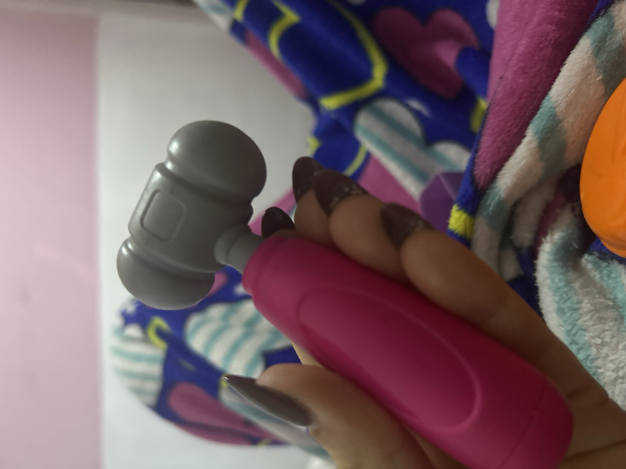 A hand with long, dark nails holds a gray and pink toy hammer. The background includes a blue, purple, and white patterned blanket, a pink wall, and an orange object. There is no information to determine if this image was taken in San Juan de Los Morros, Venezuela.