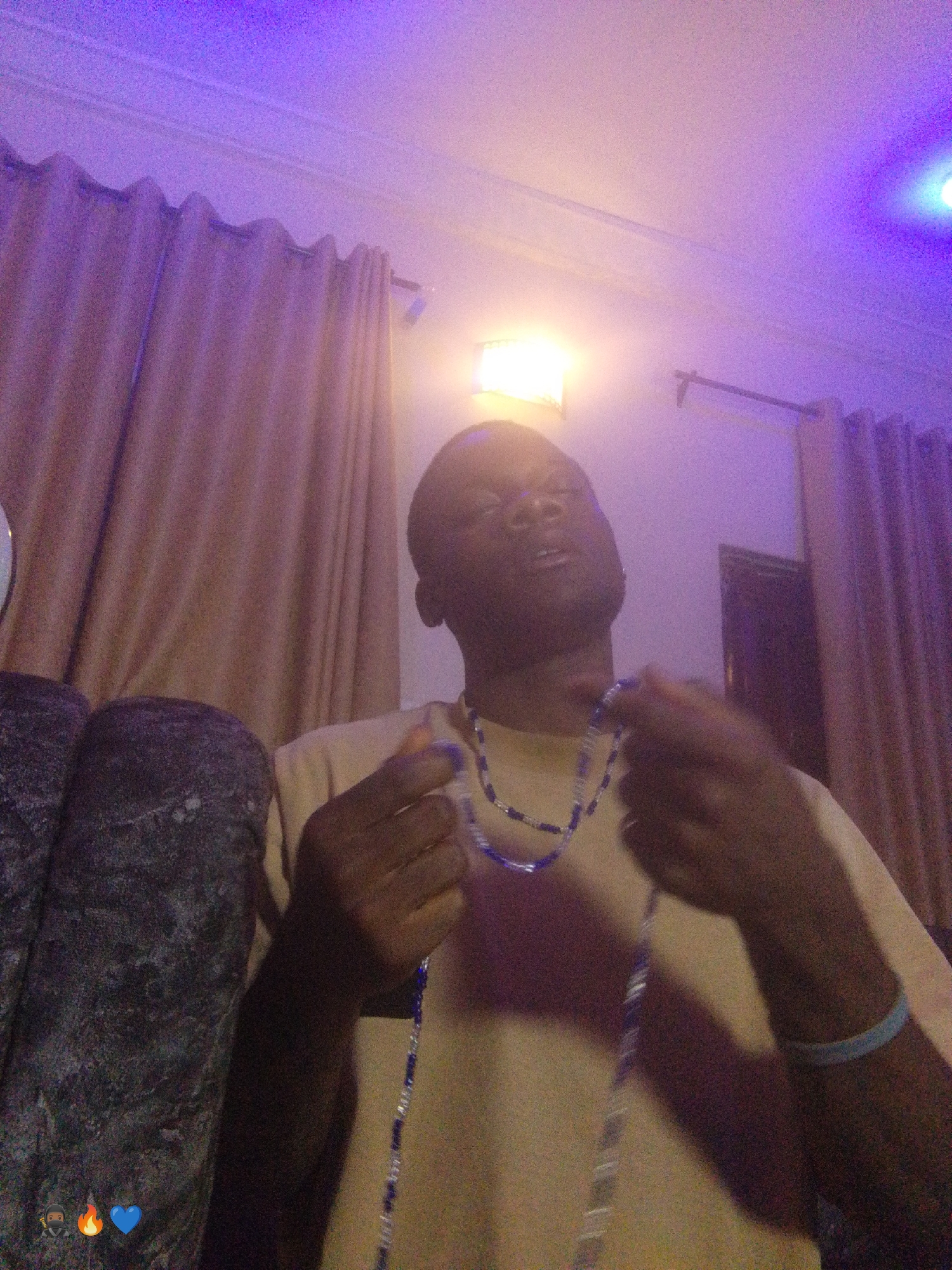 The image shows a young Black male with dark skin and short hair, wearing a light-colored t-shirt and a blue beaded necklace. He is holding another blue beaded necklace in his hands, appearing to adjust or examine it. A light blue bracelet is visible on his right wrist. The background features draped curtains, a light fixture emitting warm light, and a ceiling with purple lighting. A large, dark, textured object is visible to the left of the man. The lighting suggests an indoor setting, possibly a room in a house or a lounge area. The overall ambiance is dimly lit with pops of purple and warm light. The location is identified as Enugu, Nigeria.