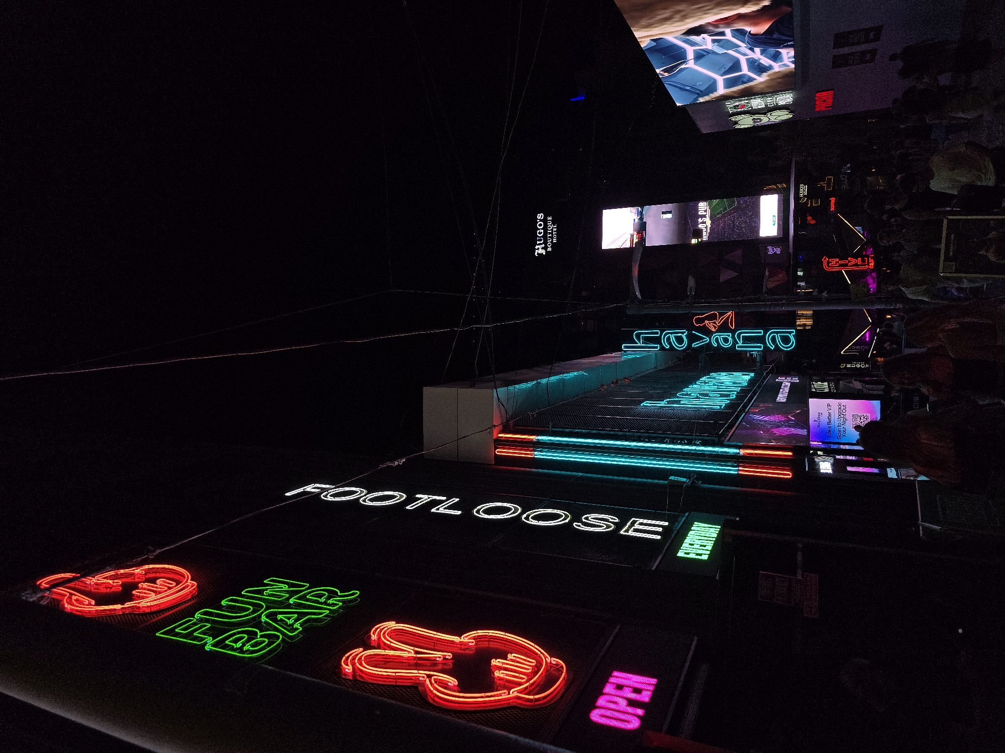 The image depicts a street scene at night in Saint Julian's, Malta, likely within the Paceville entertainment district given the prevalence of neon signs and nightlife establishments. Dominating the foreground are several neon signs, including "FOOTLOOSE" in white, and red and green neon depictions of hand gestures, possibly indicating rock-paper-scissors. To the right, a bright pink neon sign spells out "OPEN."
In the mid-ground, more neon signage is visible, including a turquoise sign that appears to read "CHAVAL" or similar. Above this, a dark building features a white neon sign that says "HUGO'S BOUTIQUE HOTEL." Various screens and other illuminated signs are also present, contributing to the vibrant, nocturnal atmosphere. The lighting is dominated by the bright colors of the neon, creating a dramatic contrast with the dark surroundings.
The overall impression is one of a lively, urban nightlife setting. The image is captured from a slightly elevated, angled perspective, looking down and across the street. The presence of reflective surfaces suggests the ground may be wet or highly polished.