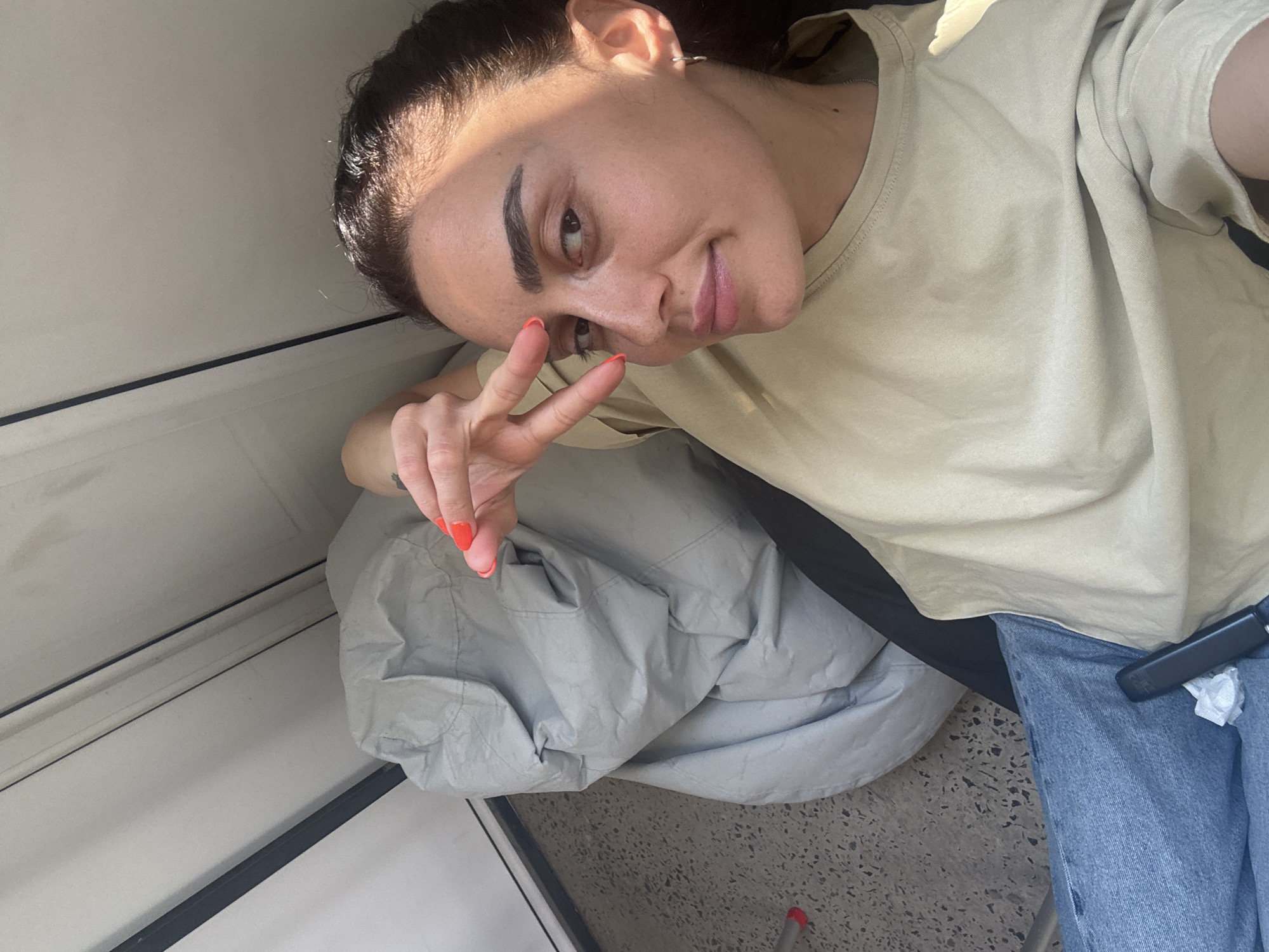 A young woman with dark hair is lying down, facing the camera, with her right hand raised to her face. She is wearing a light brown t-shirt and blue jeans. Her fingernails are painted bright orange. Next to her is a large, crumpled, light gray beanbag chair. The floor appears to be a terrazzo-style material. Behind the beanbag chair, there is a white wall or door frame. The scene is illuminated by natural light, casting shadows. The image does not provide any specific details to determine the location within the city of Tbilisi, Georgia.
