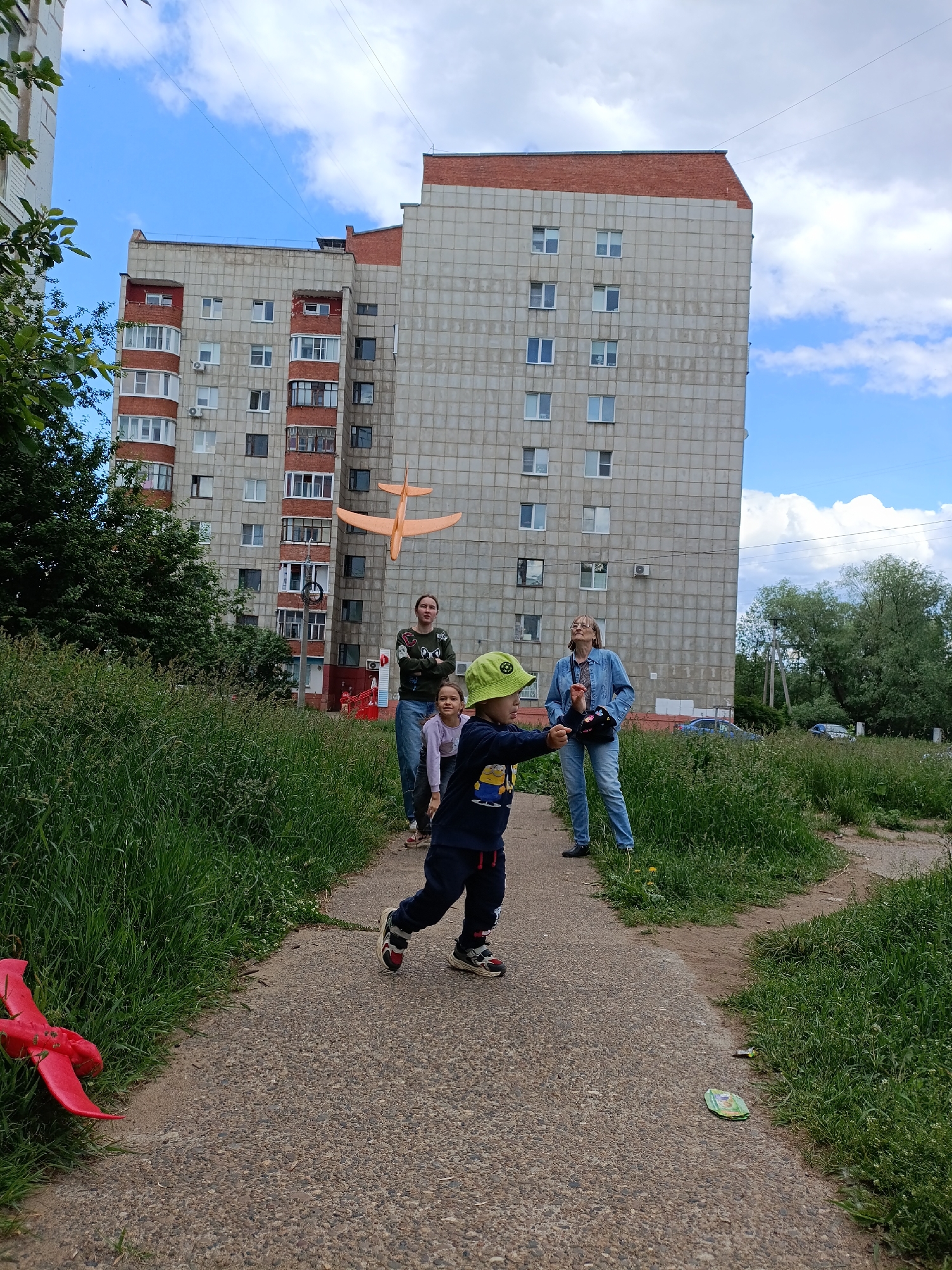 The image depicts an outdoor scene in the city of Chaikovsky, Russia, centered around a young child and several adults interacting with toy planes. In the foreground, a young boy wearing a green bucket hat, a dark blue long-sleeved shirt with a yellow graphic, and dark trousers with sneakers is positioned on a gravel path, extending his right arm forward as if in the process of throwing or having just thrown an object. An orange toy glider plane with prominent wings is suspended in mid-air behind him, slightly to the right, against the backdrop of a large residential building.
Three females are observing the scene. To the boy's left, an adult woman in a dark green sweatshirt and blue jeans stands on the path, looking towards the orange plane. A young girl in a pink top is partially visible between the boy and this woman, also looking in the same direction. Further to the right and slightly behind the boy, an older adult woman in a blue denim jacket, blue jeans, and dark shoes stands on the path, looking upwards and to the left, towards the orange plane.
The background features two multi-story residential buildings under a partly cloudy blue sky. The building immediately behind the orange plane and the observers is a large, grey concrete panel structure with numerous white-framed windows and several red brick balconies. To its left, another similar, slightly taller grey concrete building with red brick upper sections and more windows is visible. Overhead, faint power lines stretch across the sky.
The ground consists of a light-colored gravel path that forks in the middle ground, bordered by dense, overgrown green grass on both sides. To the far left, partially obscured by the grass, lies a broken red toy plane. To the far right, near the path's edge, a small green disc-like object rests in the grass. Beyond the path and buildings, a line of trees and distant cars can be seen under the expansive sky.