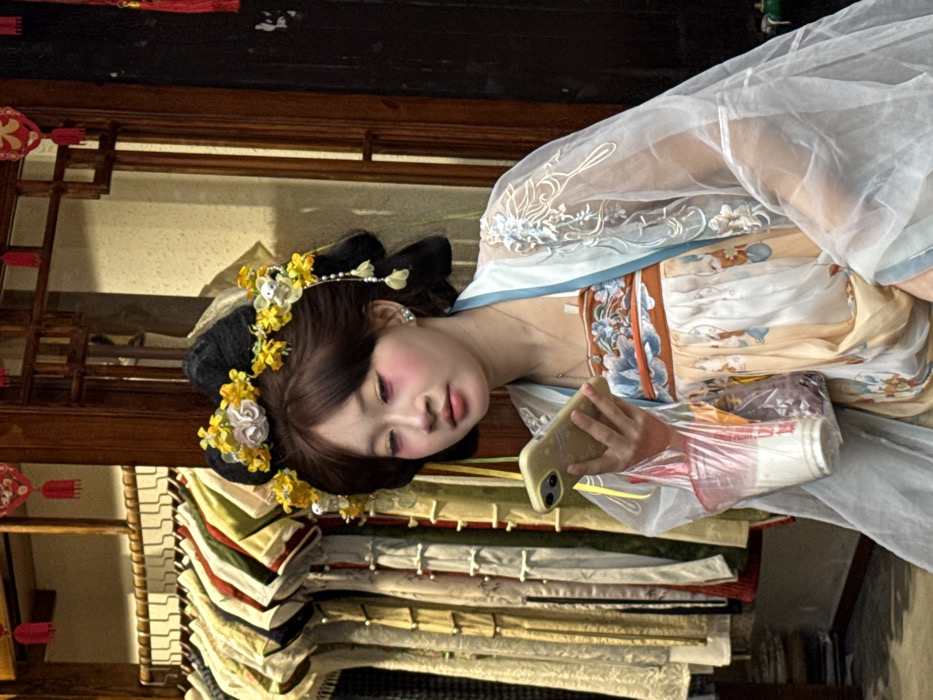 A young East Asian female is depicted in a three-quarter shot, positioned slightly right of center, gazing downwards at a light-colored smartphone held in her left hand. She wears a traditional Chinese Hanfu ensemble, consisting of an inner garment with blue and orange floral patterns, and a sheer, light blue outer robe with white embroidery. Her dark hair is arranged in a traditional high bun adorned with yellow and white floral accessories and beaded drops. Her face exhibits prominent pink cheek blush and eyeshadow. Below the smartphone, she also holds a clear plastic bag containing two plastic beverage cups, one white and one dark-colored. The background to her left features a clothing rack displaying multiple traditional garments in various colors. To the right, dark wooden lattice window frames and red decorative lanterns are visible. The environment, characterized by traditional architecture and attire, indicates a cultural tourist or commercial street in Shapingba District, China, consistent with locations such as Ciqikou Ancient Town.
