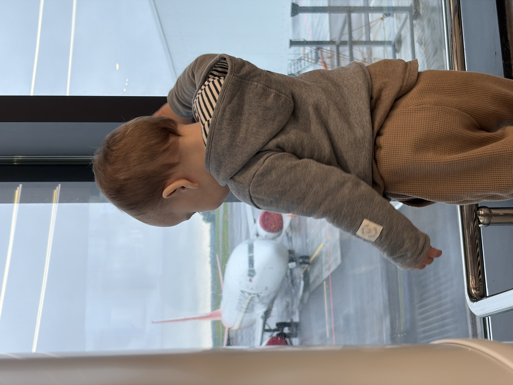 A young child wearing a gray hooded sweatshirt and brown pants is looking out of a window at an airplane on a tarmac. The child is positioned near a chrome railing. The location appears to be inside a terminal building. The city of Новое Мартьяново, Russia, is not discernible from the image.