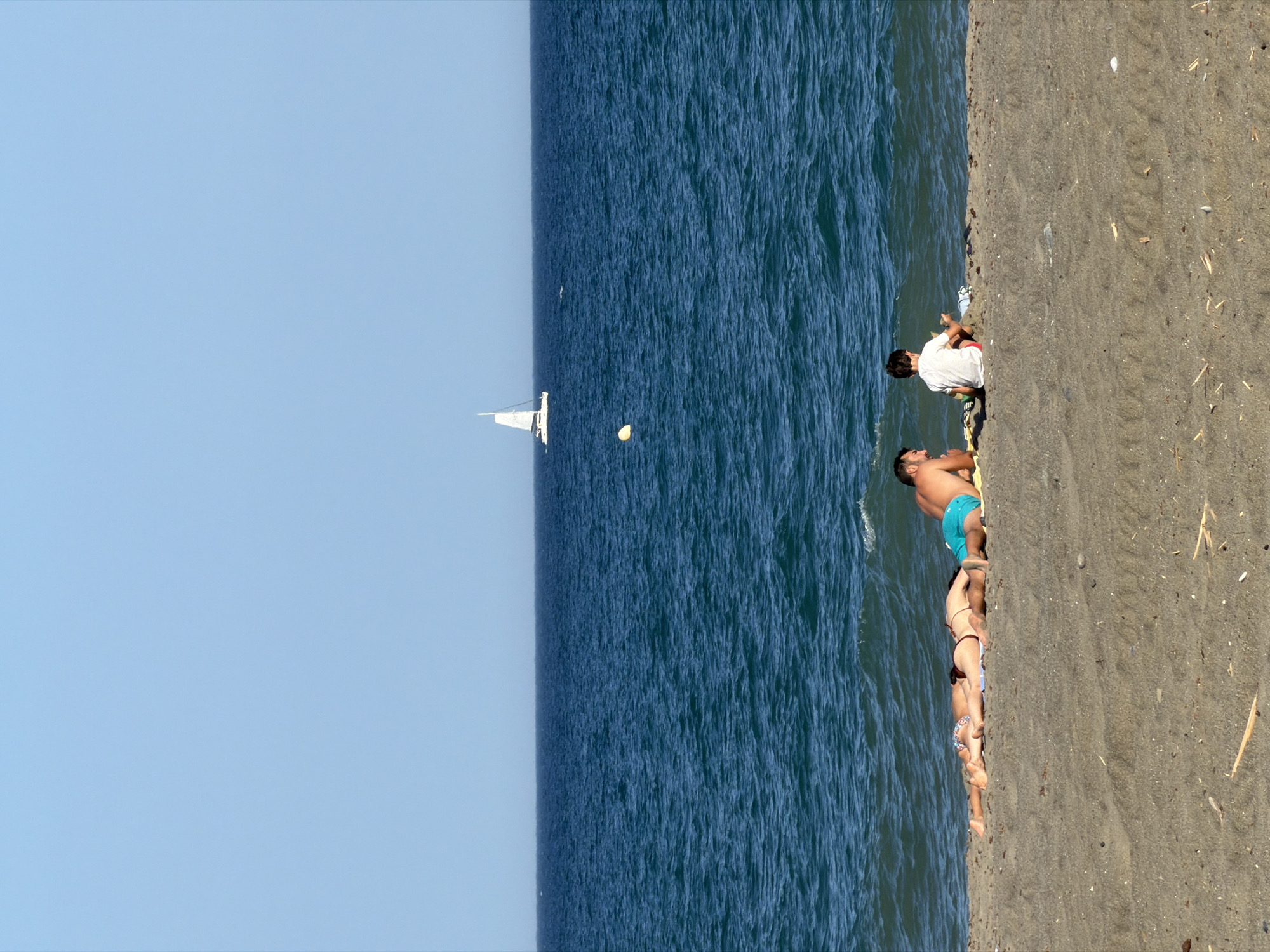 The image depicts a serene beach scene on a sunny day in Estepona, Spain. In the foreground, four individuals are visible on the sandy shore. Closest to the viewer, three people are lying down on beach towels. Two individuals are lying on their stomachs, one facing left and the other facing right, wearing bikinis. A third person, presumably an adult male, is lying on his back with his head resting on a yellow towel, wearing turquoise swimming trunks. To the right of this group, a young boy is seated, facing the ocean, wearing a white t-shirt and red shorts, appearing to play with sand or objects.
The middle ground of the image is dominated by the deep blue, slightly choppy waters of the Mediterranean Sea. A white sailboat with its sails up is visible in the distance, sailing on the horizon. A yellow buoy floats in the sea closer to the shore. The horizon line is clearly defined against a pale blue sky, suggesting clear weather. The beach itself is composed of dark, fine sand with scattered dried plant debris. The overall atmosphere is one of relaxation and leisure, characteristic of a coastal resort town.