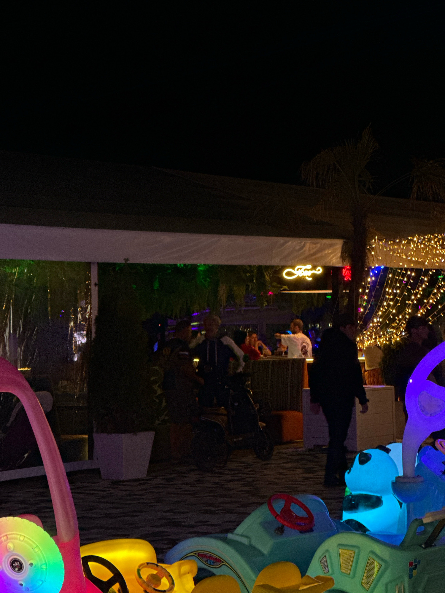 The image depicts an outdoor evening scene at what appears to be a restaurant or entertainment venue in Lermontovo, Russia. The foreground is occupied by brightly colored toy vehicles, likely for children, with illuminated wheels and features. In the mid-ground, several people are visible, some standing and others seated. One person is riding a motorized scooter. Behind them, a structure with a white awning and translucent walls is visible, adorned with numerous small, yellow lights. A neon sign that reads "Fray" in a cursive font is mounted on the awning. Palm trees are present in the background. The ground is paved with dark, patterned tiles. The overall atmosphere suggests a lively, illuminated nighttime setting.