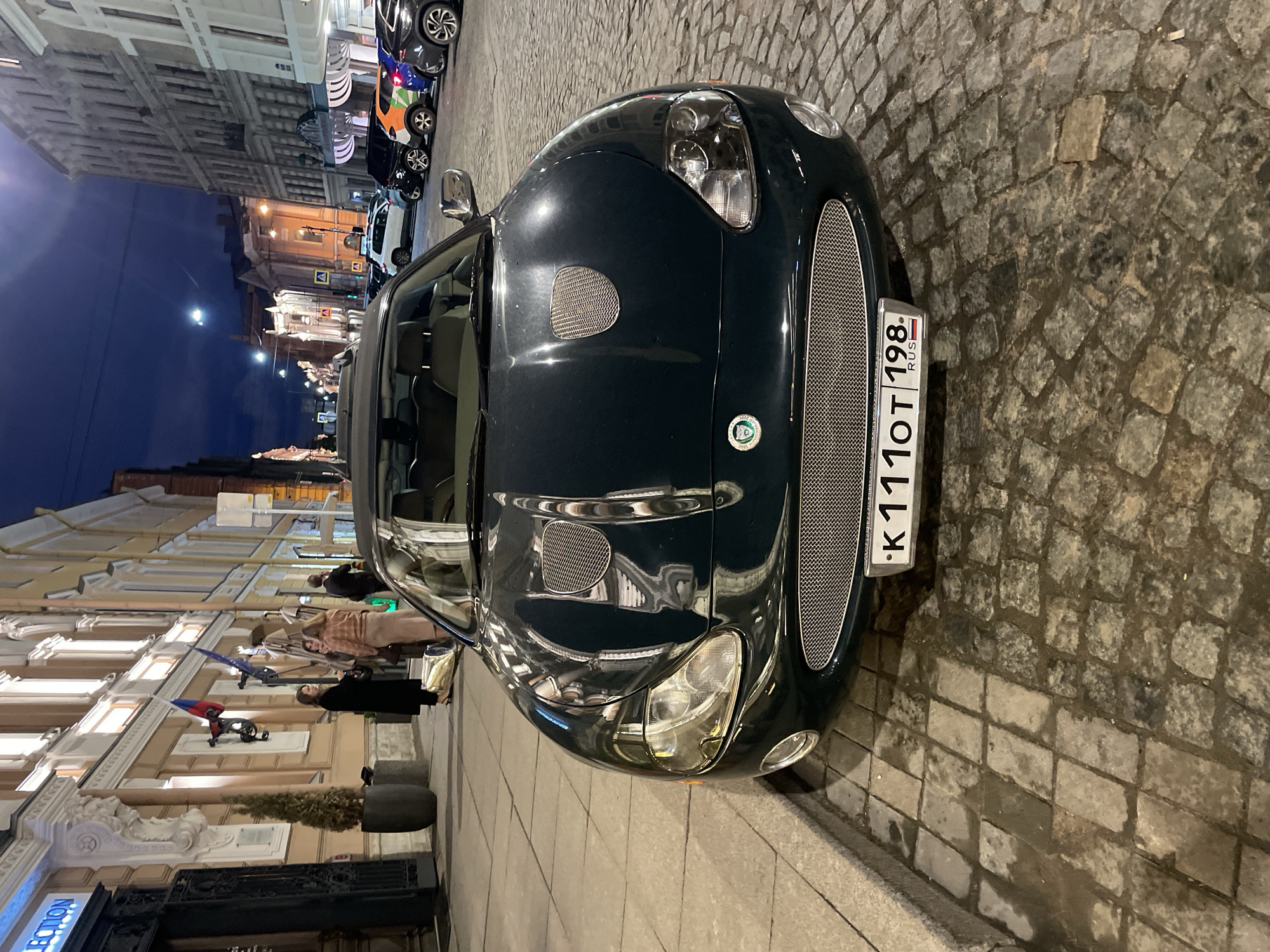 The image depicts a dark green Jaguar XK150 parked on a cobblestone street in Saint Petersburg, Russia. The car is positioned at an angle, facing towards the right of the frame. Its front grille and Russian license plate are clearly visible. The license plate reads "K111OT 198 RUS."
In the background, on the left side of the frame, are buildings with classical architectural details, including visible windows, balconies, and decorative elements. Pedestrians are visible walking along the sidewalk.
Additional cars are parked further down the street, and streetlights illuminate the scene, creating reflections on the car's hood. The sky is dusky, indicating that the photo was taken either at dusk or in the evening. The ground is comprised of unevenly sized cobblestones.