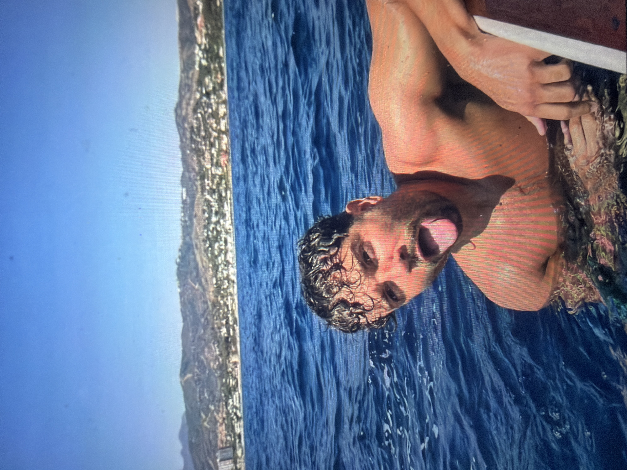 An adult male is pictured partially submerged in deep blue, choppy water, leaning against the edge of a boat or float. He has wet, dark, curly hair and stubble, and looks directly at the camera with his mouth open and tongue extended in a playful expression. His tanned torso is visible above the water, showing vertical light and shadow patterns. In the background, a distant coastline features rolling hills covered in sparse vegetation, with numerous white and light-colored buildings scattered across them, indicating a populated urban area. A taller, multi-story building is visible on the far left of the distant shore. The sky above is clear and bright blue. The scene is located in the city of Shumen, Bulgaria.