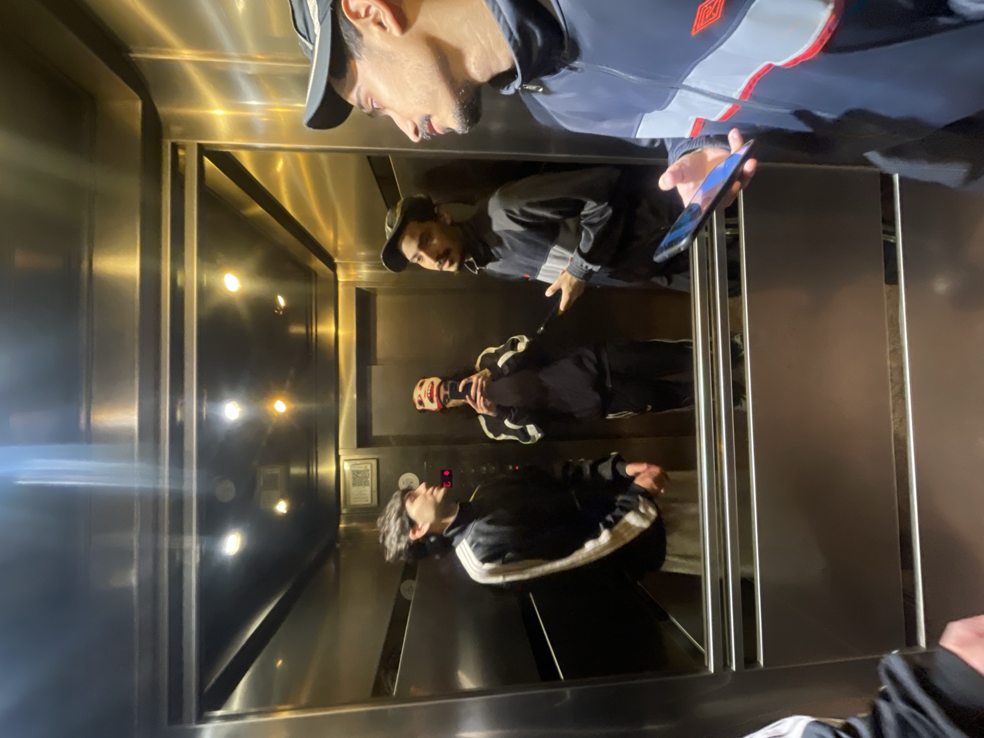 This image captures multiple individuals inside a highly reflective metal elevator, presented with a vertical orientation where the elevator's ceiling is on the left and the floor is on the right.
In the upper right portion of the frame, a man wearing a dark baseball cap and a dark blue and grey jacket with a red and white geometric logo (resembling Umbro's) is prominent. He has a beard and is holding a smartphone in his right hand, its screen showing blue and purple light, and his gaze is directed downwards.
Reflected in the shiny metallic walls across the center of the frame are two other individuals. One figure, centrally positioned in the reflection, wears a dark cap and a dark jacket with white stripes. Further to the left within the reflections, another person in a black and white striped jacket is visible, looking slightly upwards. Additionally, a faint reflection of a fourth person wearing a red and white mask or balaclava and holding a phone can be seen near what appears to be the elevator control panel.
The elevator interior is illuminated by several bright, warm-toned lights, casting strong reflections on the polished walls. An elevator control panel, located towards the lower left in the reflections, displays red digital numbers (possibly '13') and has a small, unreadable sign or QR code next to it. All individuals are dressed in casual or sporty attire. The scene conveys a moment of transit within the elevator. The location is specified as Buenos Aires, Argentina.