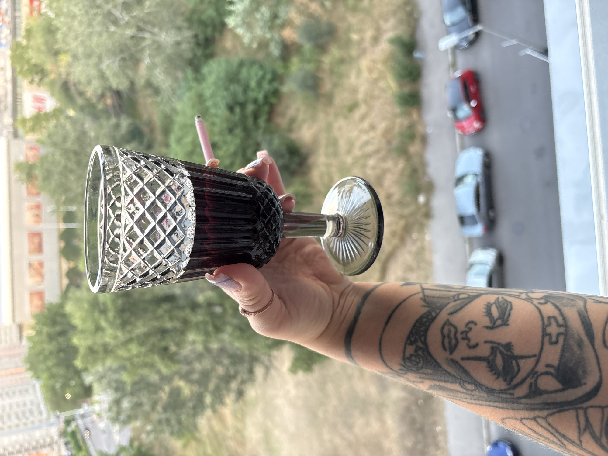 A close-up shot focuses on a person's forearm with a prominent tattoo of a face. The arm is holding a stemmed glass filled with dark red wine. A lit cigarette with a pink filter is held between two fingers, resting near the rim of the glass. In the background, a blurry cityscape unfolds, with greenery and buildings visible. The blurred image of cars parked on a street is discernible in the mid-ground, and a building with "H&M" signage can be seen in the distance, suggesting an urban environment, possibly Belgrade, Serbia.