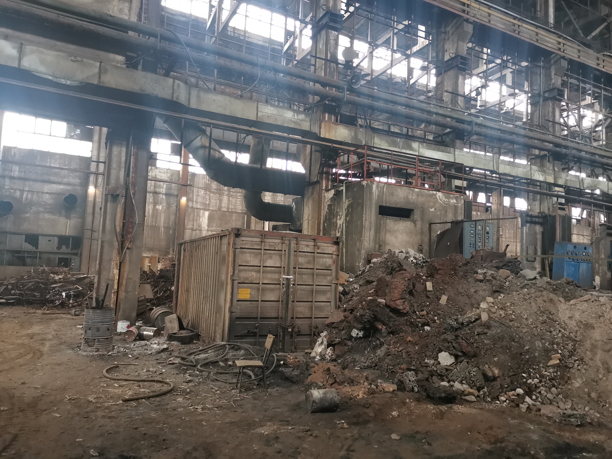 The image depicts an interior space, seemingly a factory or industrial site, possibly located in Zhivkovo, Bulgaria. The environment is characterized by a dirt floor, scattered debris including metal scraps, cables, and barrels, and a large pile of what appears to be earth and rock. An intermodal container sits adjacent to the pile. The background consists of concrete pillars and beams, industrial piping, and rows of windows suggesting a dilapidated state. No people are visible.