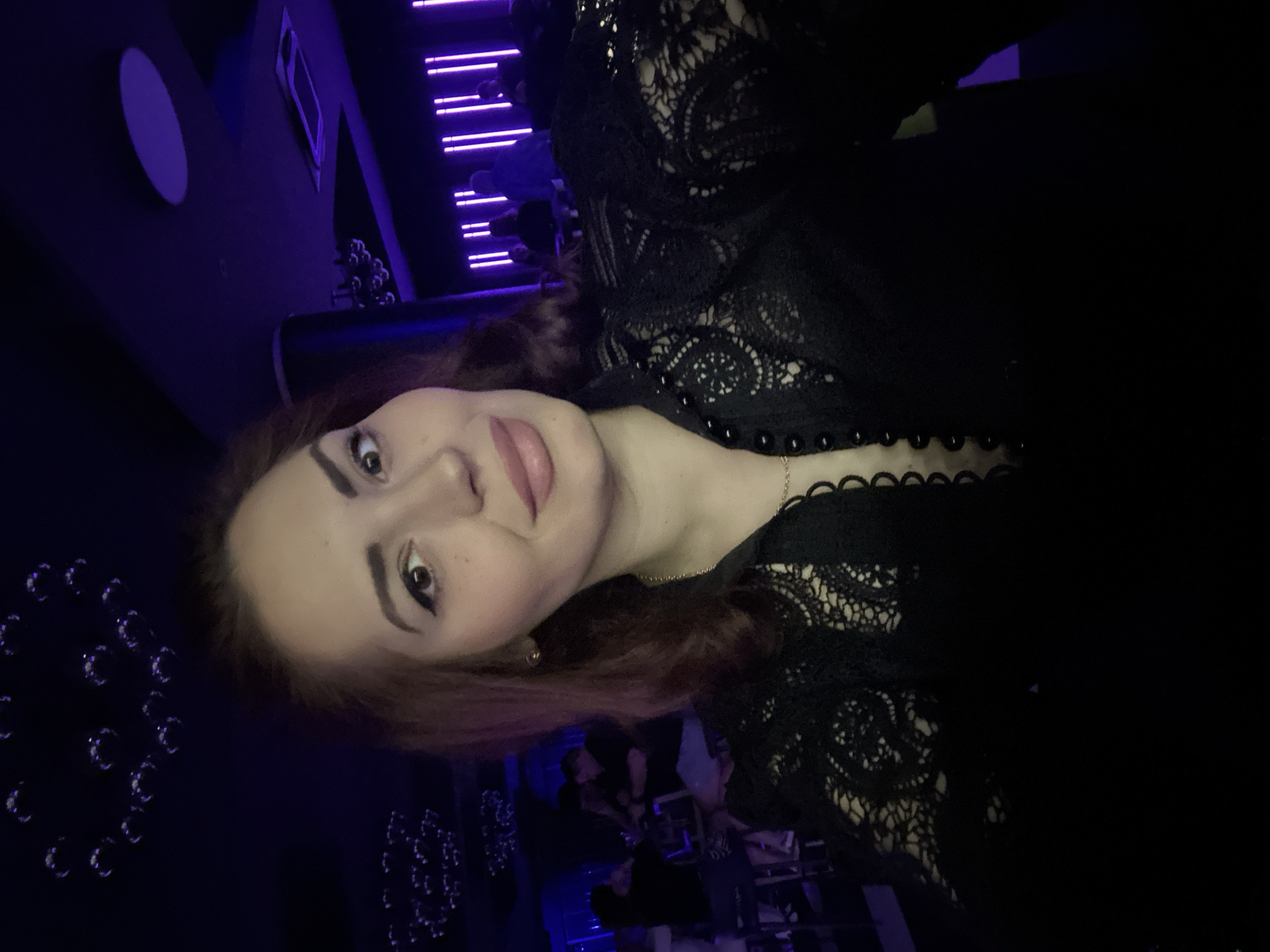 The image displays a young woman in a dimly lit, modern social venue, likely a bar or club, at night in Kazanlak, Bulgaria. She is captured in a close-up, looking directly at the camera with a neutral to slightly pleasant expression. Her reddish-brown hair is visible, and her face features eyeliner and pink lipstick. She is wearing a black top with intricate lace patterns and a scalloped neckline adorned with small black beads, complemented by a thin gold chain necklace.
The background is characterized by deep blue and purple ambient lighting, creating a dark, nocturnal atmosphere. Visible decorative elements include a large circular light fixture on a dark wall, as well as patterns of smaller, shiny circular ornaments. In the upper right background, several parallel, thin purple LED lights run horizontally, suggesting a contemporary design. Faint, blurry figures of other people and outlines of tables are discernible in the deeper background, indicating a social gathering or event. The scene evokes a trendy, indoor nightlife setting. No text is visible in the image.