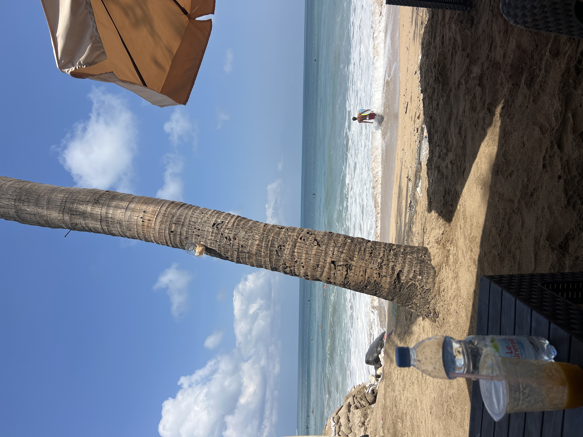 The image depicts a sunny day at Kuta Beach, Indonesia, with a view of the ocean. In the foreground, a dark table holds a clear plastic cup with a straw and an amber-colored drink, and a plastic bottle of water. To the left of the table, sandbags and rocks line the edge of the beach. A large palm tree trunk is prominent on the left side of the frame. In the background, the turquoise ocean meets the shore with white waves crashing. Several surfers are visible in the water. A person in red shorts and holding a blue and yellow surfboard is walking into the ocean. An umbrella is partially visible in the upper right corner of the image. The sky is a clear blue with scattered white clouds.