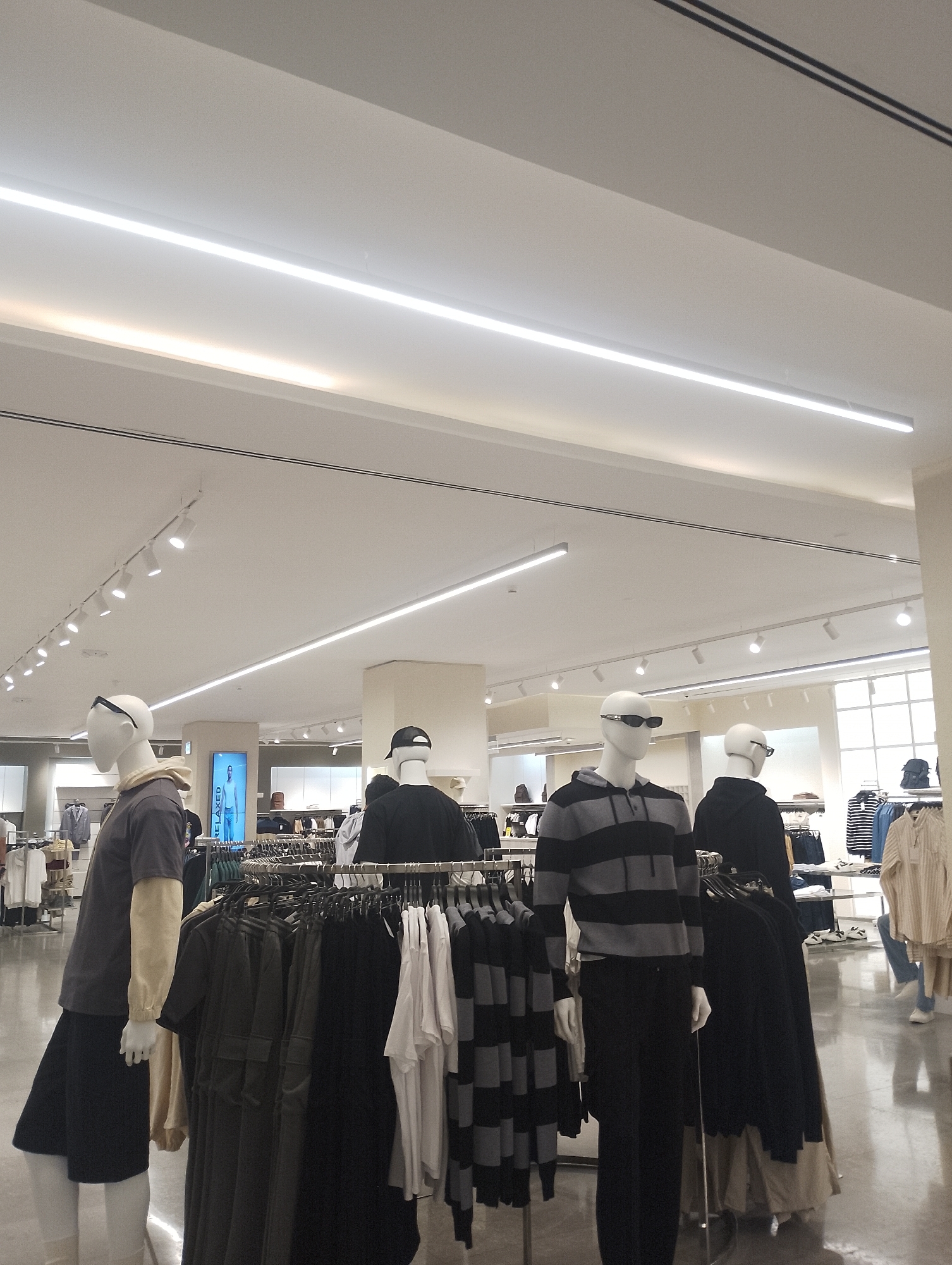 The image shows the interior of a clothing store, likely located in a shopping mall or retail complex, though the specific location in Pampatar, Venezuela, cannot be confirmed from the image alone. The scene is illuminated by fluorescent ceiling lights. Several mannequins are on display, dressed in casual clothing like hoodies, t-shirts, and pants. Racks of clothes surround the mannequins, filled with items such as black shirts, sweaters, and striped tops. In the background, other clothing racks are visible, suggesting a wide selection of apparel is available. The floor is shiny and reflective, likely tiled or polished. There are no customers currently in the immediate foreground of the image.