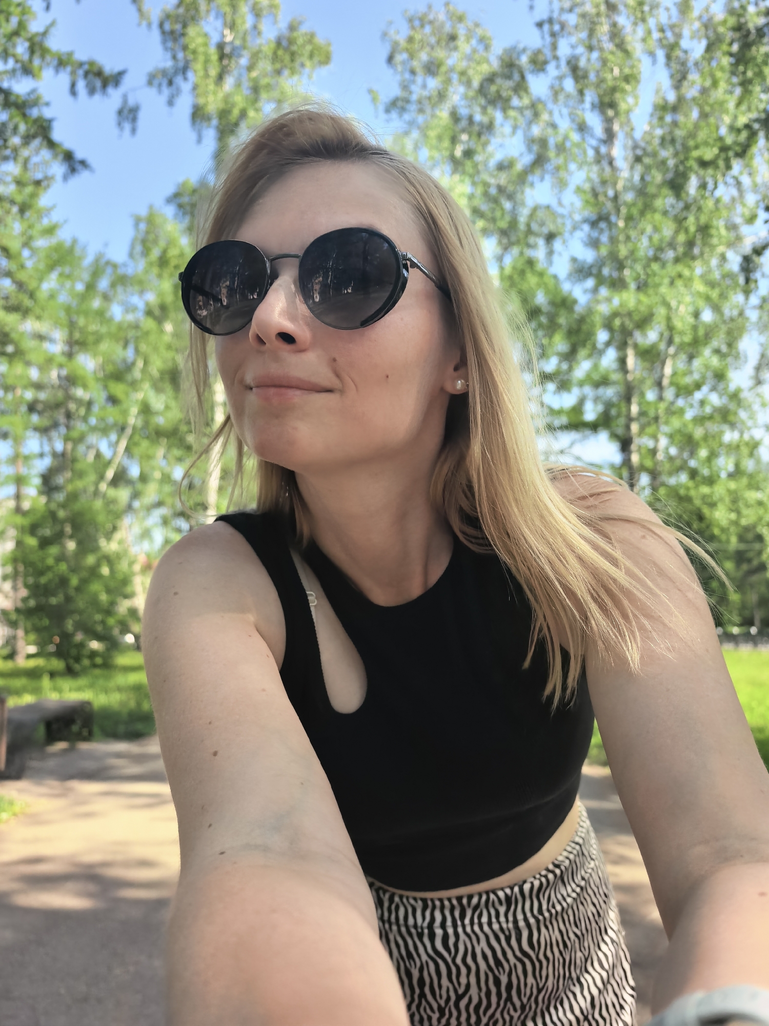 A woman is taking a selfie outdoors, likely in a park. She has blonde hair and is wearing round black sunglasses, a black top with a cutout, and a zebra-print skirt. She is positioned in front of a background of green trees and a blue sky. The trees are illuminated by sunlight. In the distance, a bench is visible on the left side. The location is likely in a park in Sterlitamak, Russia, as specified by the prompt.