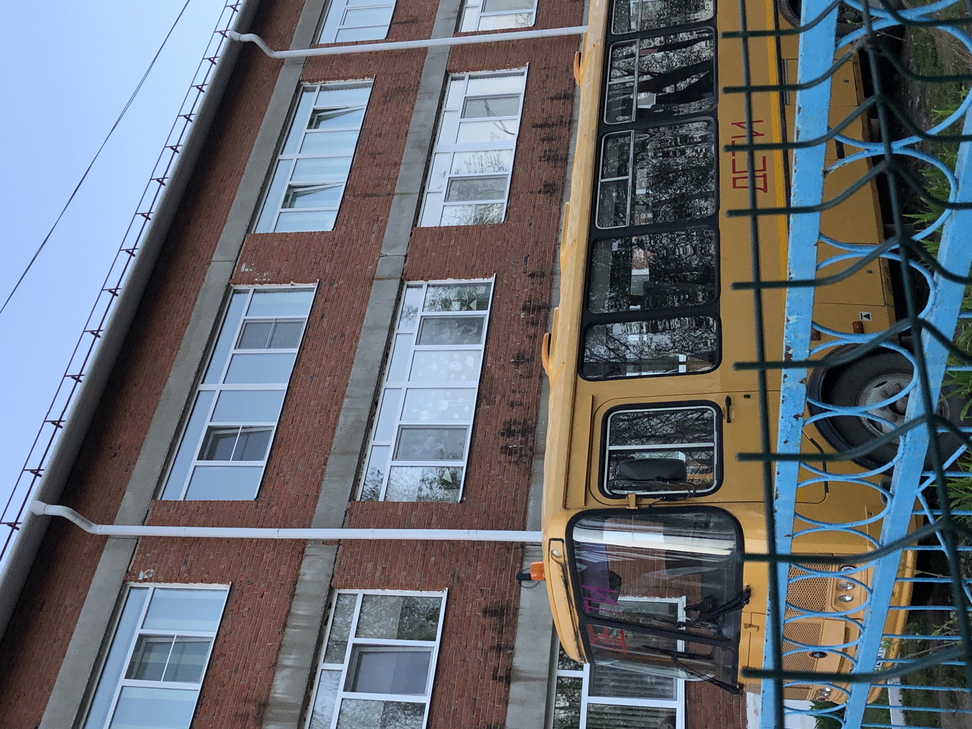 The image shows a multi-story brick building next to a yellow bus, partially obscured by a blue metal fence. The building has multiple rows of windows. The bus has the word "ДЕТИ" (children) written on its side. The location appears to be in Russia, given the Cyrillic text.