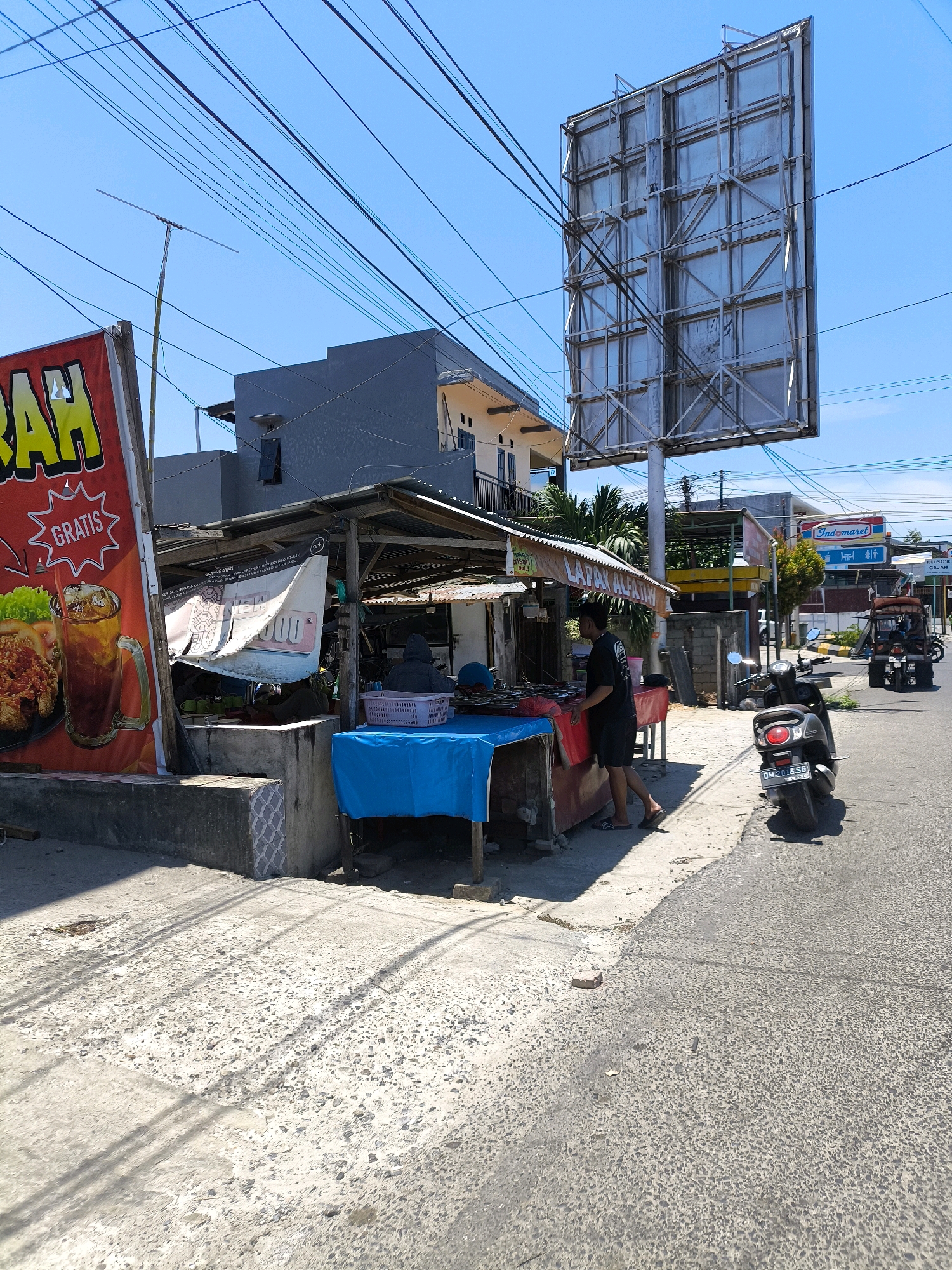 This is a street-level photograph taken in Gorontalo, Indonesia. The scene depicts a roadside stall with various items for sale, including what appears to be food. A person is standing behind the stall, and another person is seated. A blue tarp is spread over a portion of the stall. In the foreground, a black scooter is parked on the right side of the road. Further down the road, a three-wheeled vehicle is driving away. A large billboard is visible in the background, along with overhead power lines and buildings. A banner advertising "GRA TIS" (Free) and an image of food and iced tea is prominently displayed on the left side of the image. The ground is paved with asphalt and concrete, showing signs of wear and tear. The sky is clear and blue, indicating a sunny day.