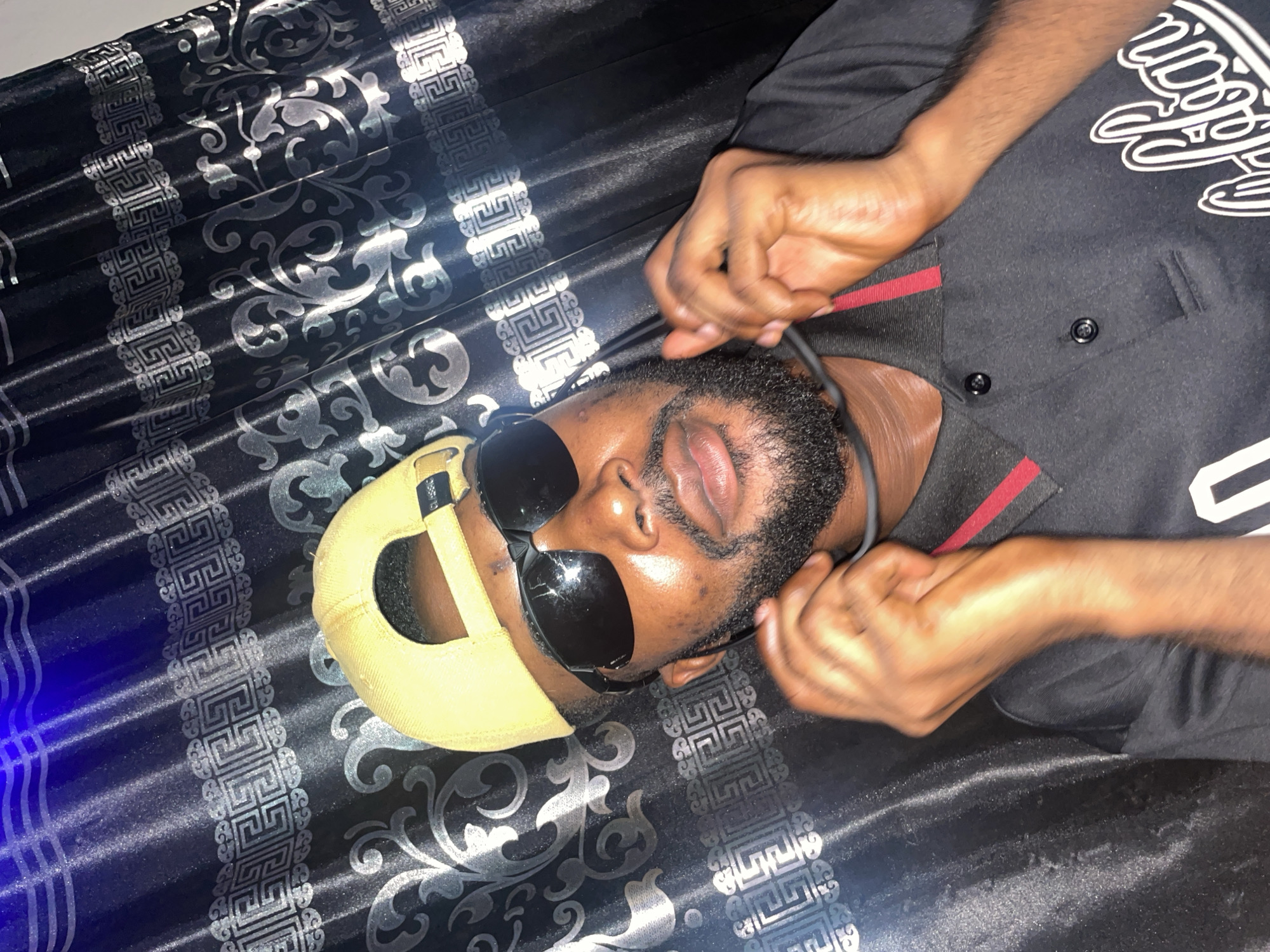 A man with a beard and wearing sunglasses and a yellow baseball cap is lying on a patterned black and silver surface. He's wearing a black shirt with white text and red trim. His hands are raised near his face. The location is not discernible in the image; therefore, identifying it as Lokoja, Nigeria, is not possible.