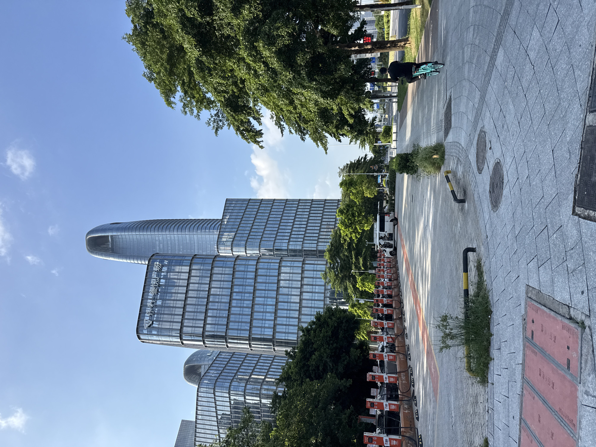 The image captures a bright, sunny day in Haizhu District, China. The scene features a modern cityscape dominated by sleek, glass-facade skyscrapers that reflect the clear blue sky and scattered white clouds. Lush green trees provide a natural contrast to the urban architecture. In the foreground, a paved walkway is lined with a row of orange and white electric scooters. A person on a bicycle, wearing dark clothing, is seen riding along the walkway, casting a shadow on the ground. The time of day appears to be late morning or early afternoon, given the angle of the sun and the length of the shadows. The overall atmosphere is one of urban vibrancy and technological advancement, with a hint of leisure suggested by the presence of the scooters and the cyclist. Visible text is present on the side of the buildings, appearing to be in Chinese characters, though it is not clearly legible.
