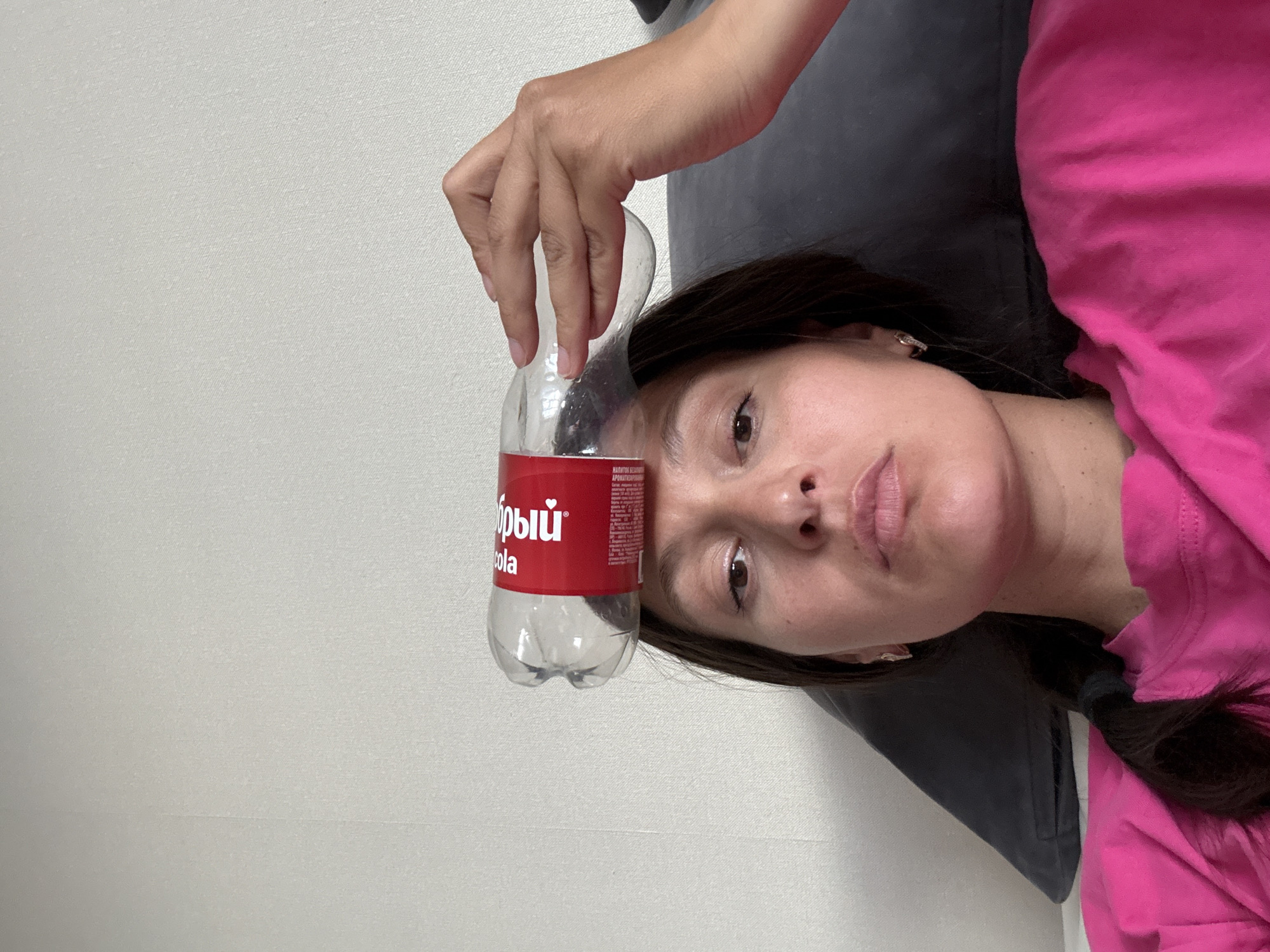 A woman lies on a dark gray couch, wearing a pink shirt. A half-full, red and clear plastic bottle labeled "Добрый cola" is held above her face. The background is a plain white wall. The location is likely Chaikovsky, Russia, based on the bottle label.