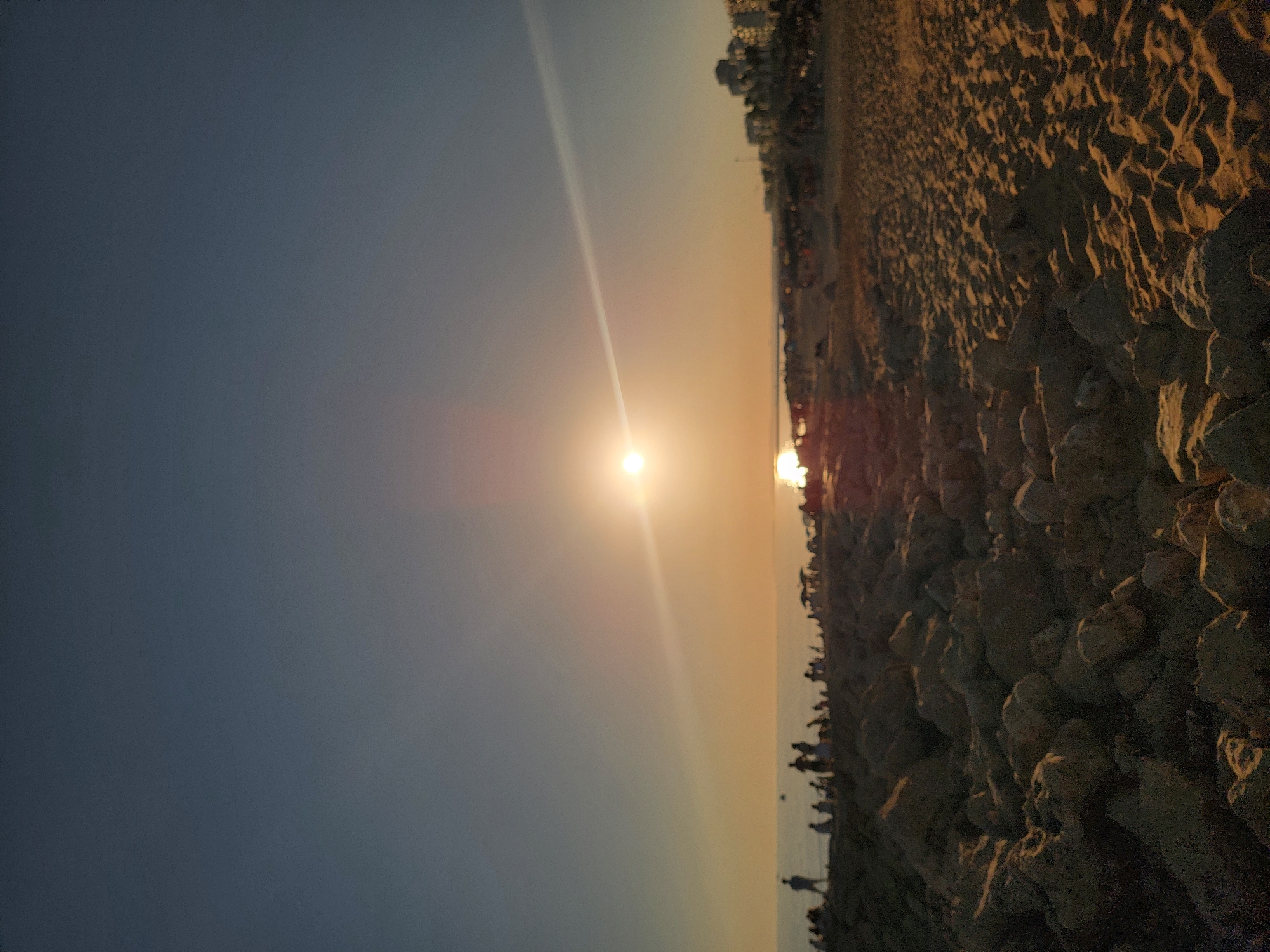 The image depicts a sunset scene at a beach, likely in Quarteira, Portugal. The sun is low on the horizon, casting a warm, golden light across the sky and illuminating the sandy beach and rocky shoreline. Silhouettes of people can be seen gathered along the water's edge, enjoying the view. The ocean is calm, reflecting the colors of the sunset. Buildings are visible in the background, suggesting a developed coastal area. The overall mood is serene and peaceful, capturing a moment of natural beauty and shared human experience.