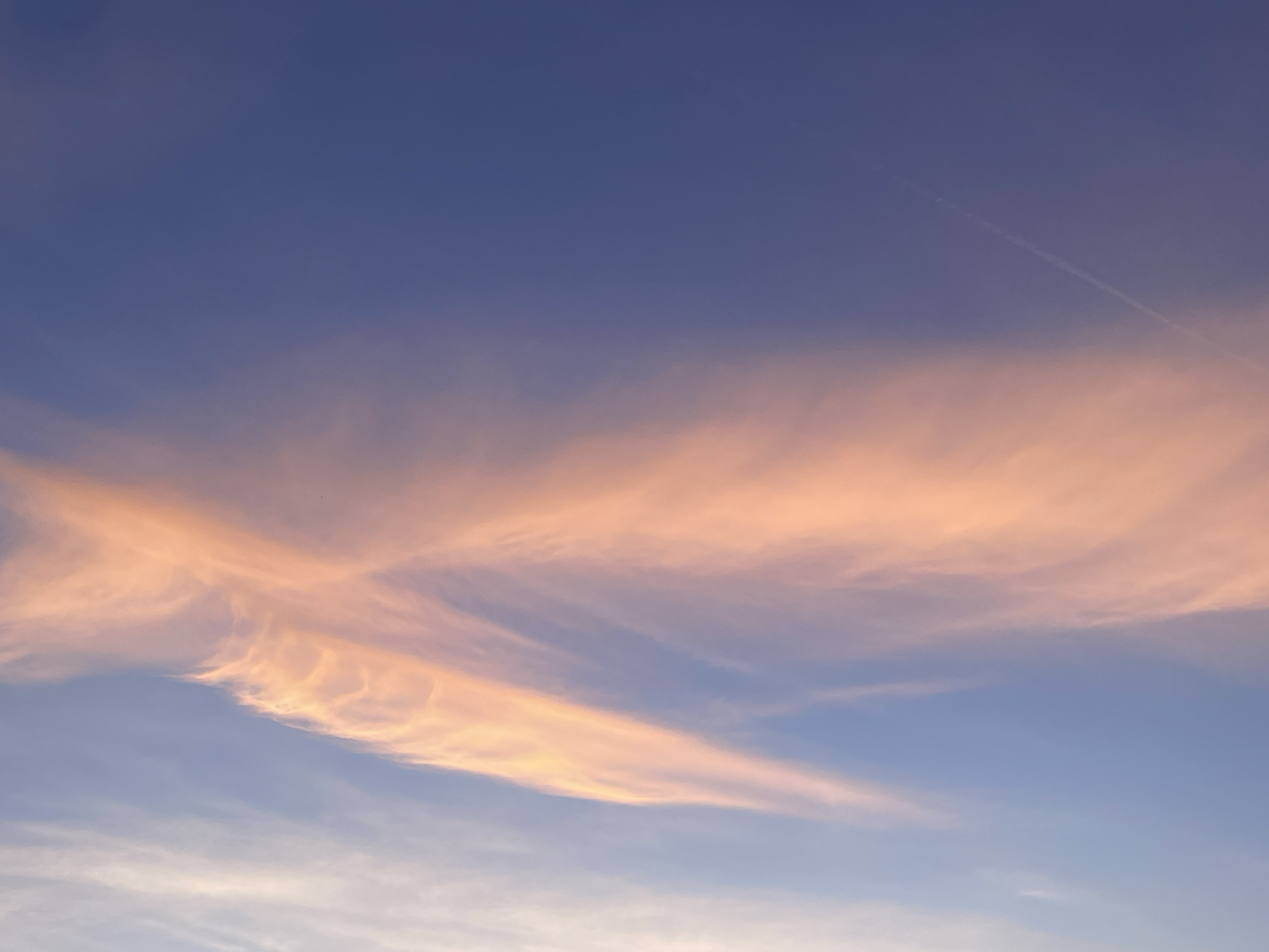 The image captures an expansive view of the sky over Bar, Montenegro. The upper portion of the sky presents a deep purplish-blue hue, gradually transitioning to a lighter blue-grey closer to the bottom. Elongated, wispy cloud formations, illuminated in shades of orange and pink, are prominently visible across the central and upper-central sections of the frame. A faint, thin white contrail is discernible, traversing diagonally through the upper-right quadrant of the sky. No people, discernible subjects, objects, actions, or interactions are present within the scene.