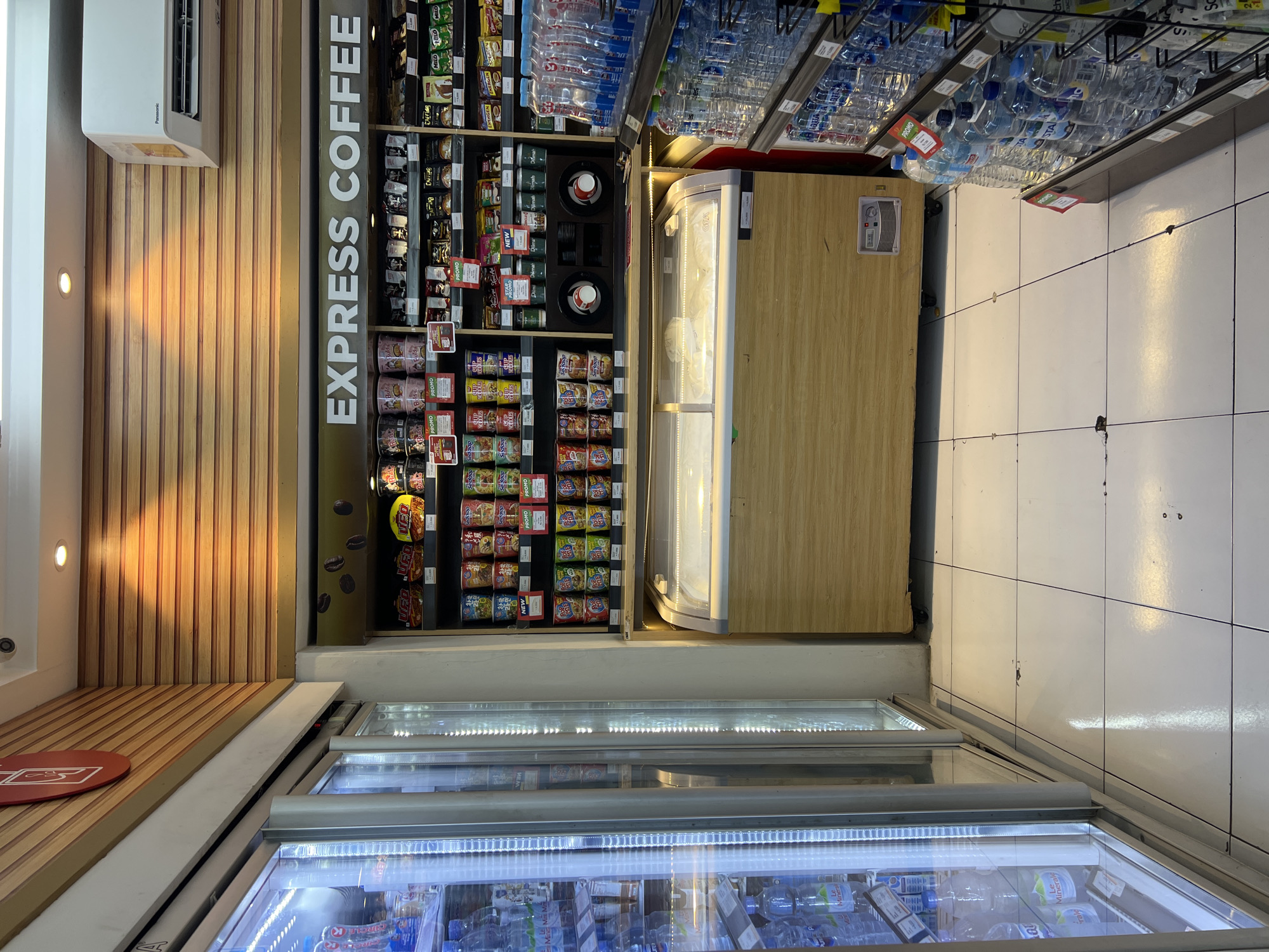This is an indoor shot of a convenience store in Denpasar, Indonesia. The store features a large refrigerated display case on the left filled with bottled drinks. To the right, shelves are stocked with various food and beverage items, including instant noodles and coffee. Above the shelves, "EXPRESS COFFEE" is written in large white letters. A wooden-fronted freezer unit is positioned in the center of the image. The floor is tiled in white. An air conditioning unit is mounted on the wall above the shelving unit. The scene appears to be empty of customers.