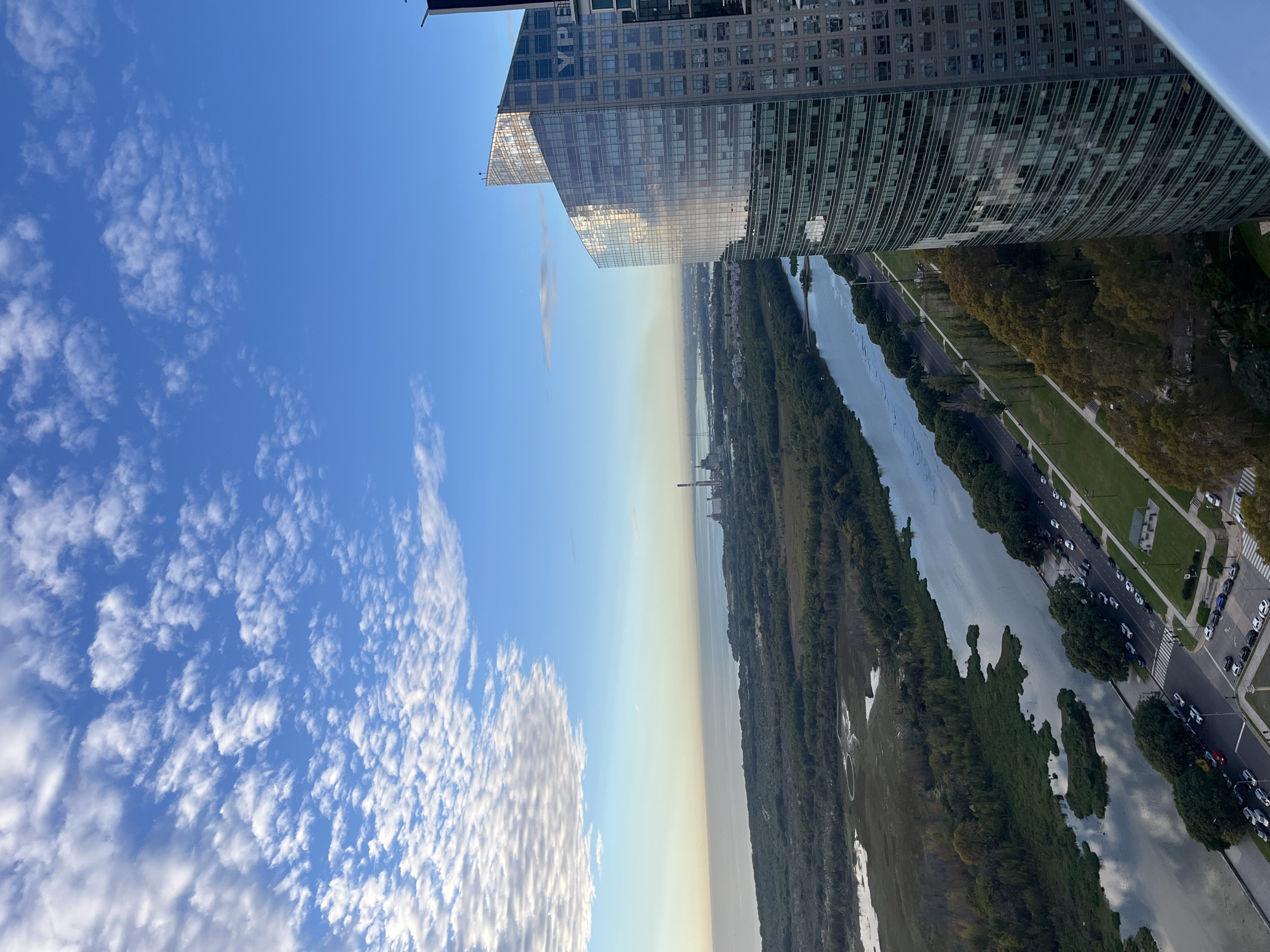 The image depicts a cityscape with a bright blue sky filled with scattered clouds. The top of two tall modern buildings with reflective glass facades dominates the upper part of the frame. Below the buildings, a wide body of water, possibly a river, is visible, with a dense strip of greenery along its banks. A road runs parallel to the river, lined with trees and cars. In the distance, industrial structures are faintly visible.
The location is likely the Puerto Madero district in Buenos Aires, Argentina, known for its modern architecture, waterfront, and adjacent ecological reserve along the Rio de la Plata. The angle and perspective suggest the photograph was taken from a high vantage point within one of the buildings.