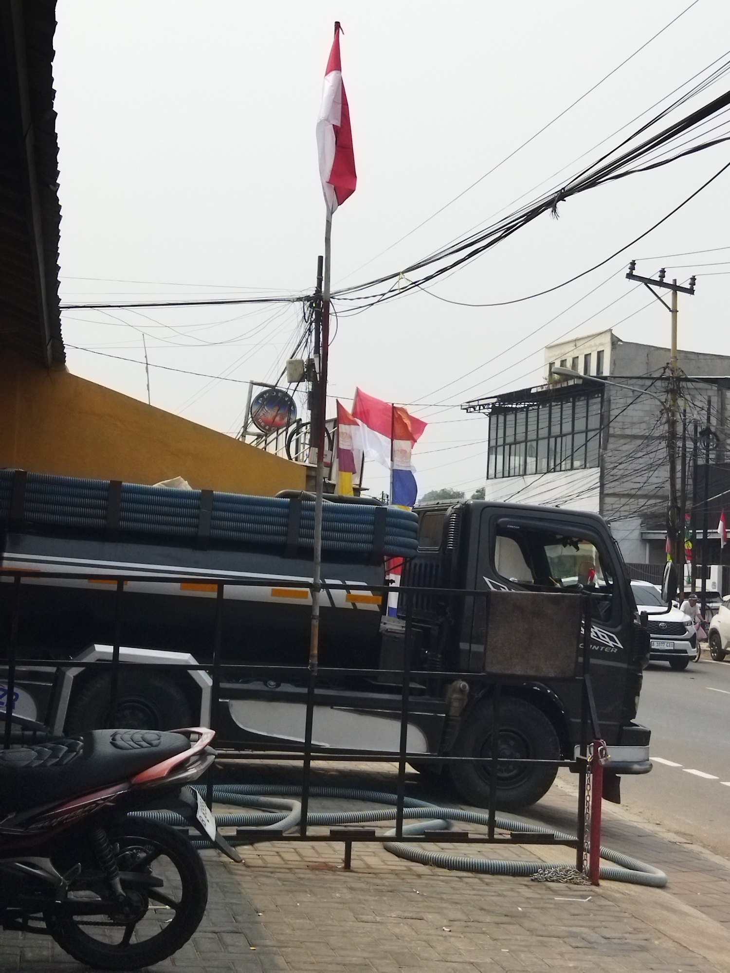 A side profile of a black truck is visible on a street in Tangerang, Indonesia. The truck is loaded with blue corrugated pipes. In front of the truck, a motorcycle is parked. Indonesian flags are displayed on a pole. The street is paved with bricks and there are electrical wires overhead. In the background, there are buildings and other vehicles. The sky is overcast.
