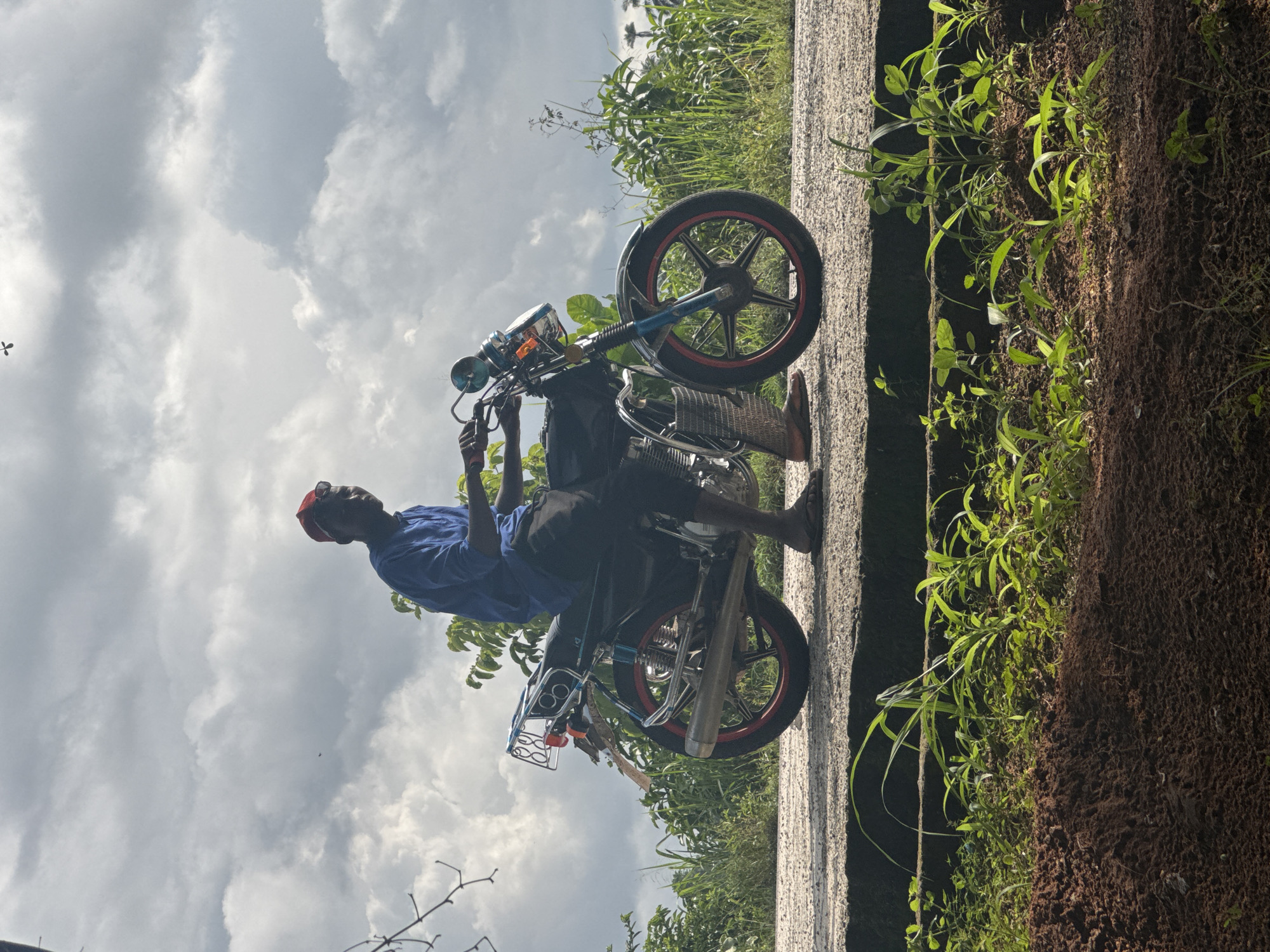A man sits on a motorcycle on the side of a road in Ihiagwa, Nigeria. The motorcycle has red rims. The road is concrete. Weeds and plants grow on the side of the road. The sky is cloudy.