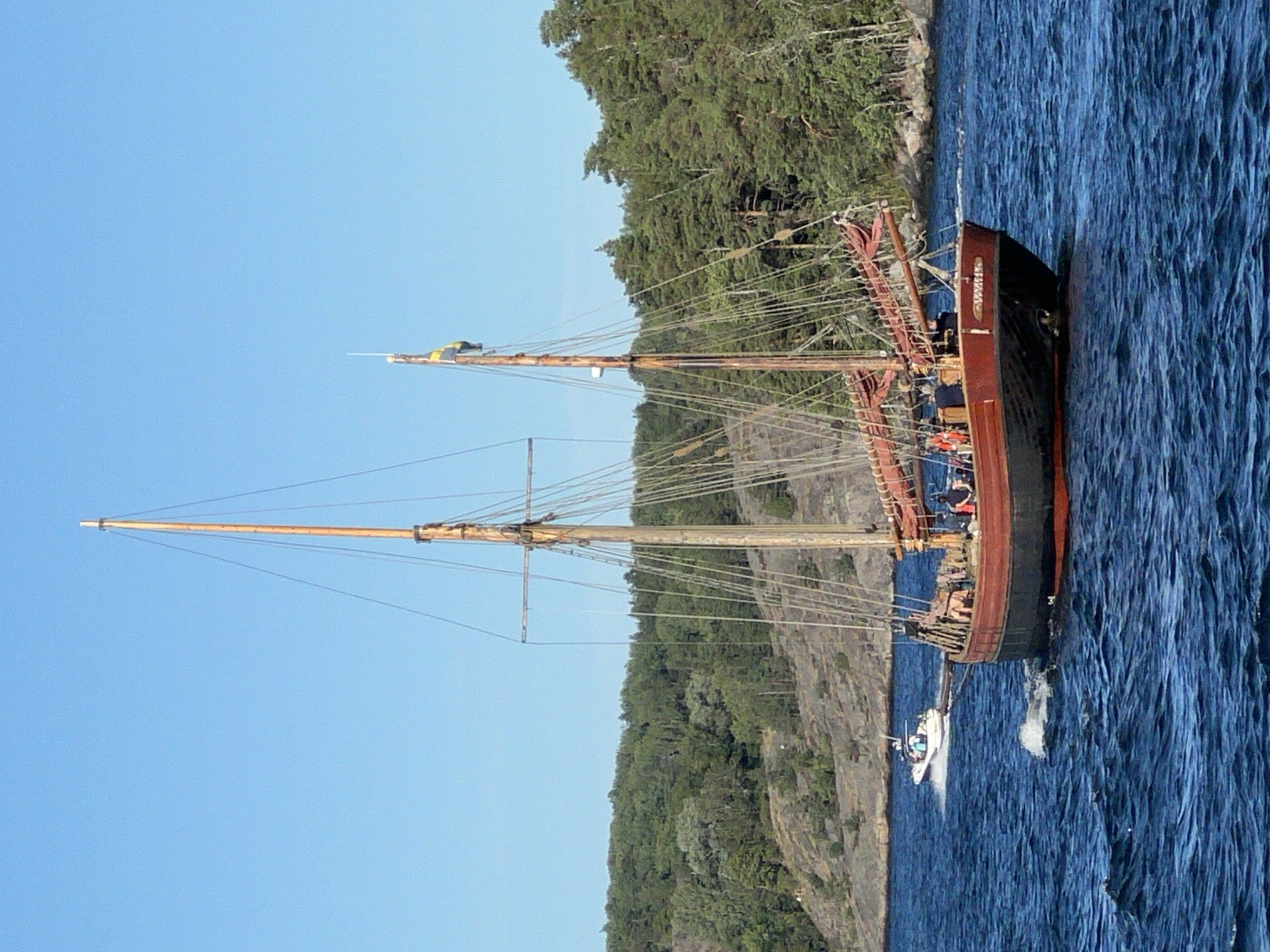 A wooden sailing ship with two masts is sailing on a body of water. The ship is red and black and has several people on deck. There is a smaller boat in the background. The ship is likely in the archipelago near Nacka, Sweden, given the presence of rocky islands covered with trees. The water is blue and choppy. The sky is clear and blue. The overall scene suggests a pleasant day for sailing.