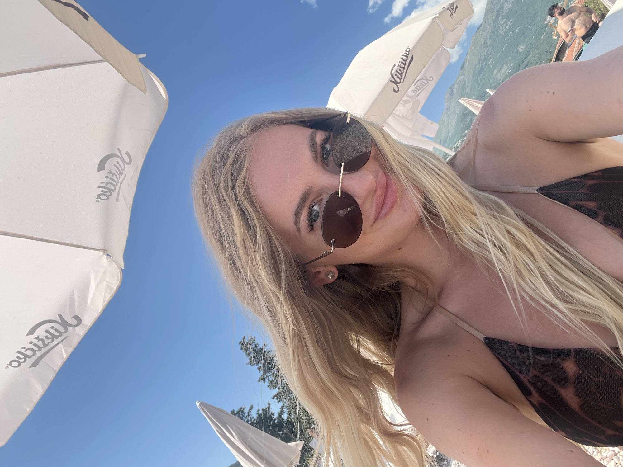 A woman is taking a selfie at the beach in Budva, Montenegro. She is wearing a brown and black patterned bikini and sunglasses, and has long blonde hair. She is standing under a white beach umbrella with the word "Nikšičko" printed on it. In the background, there are mountains and another beach umbrella. A man is visible in the background. The sky is blue with some clouds.