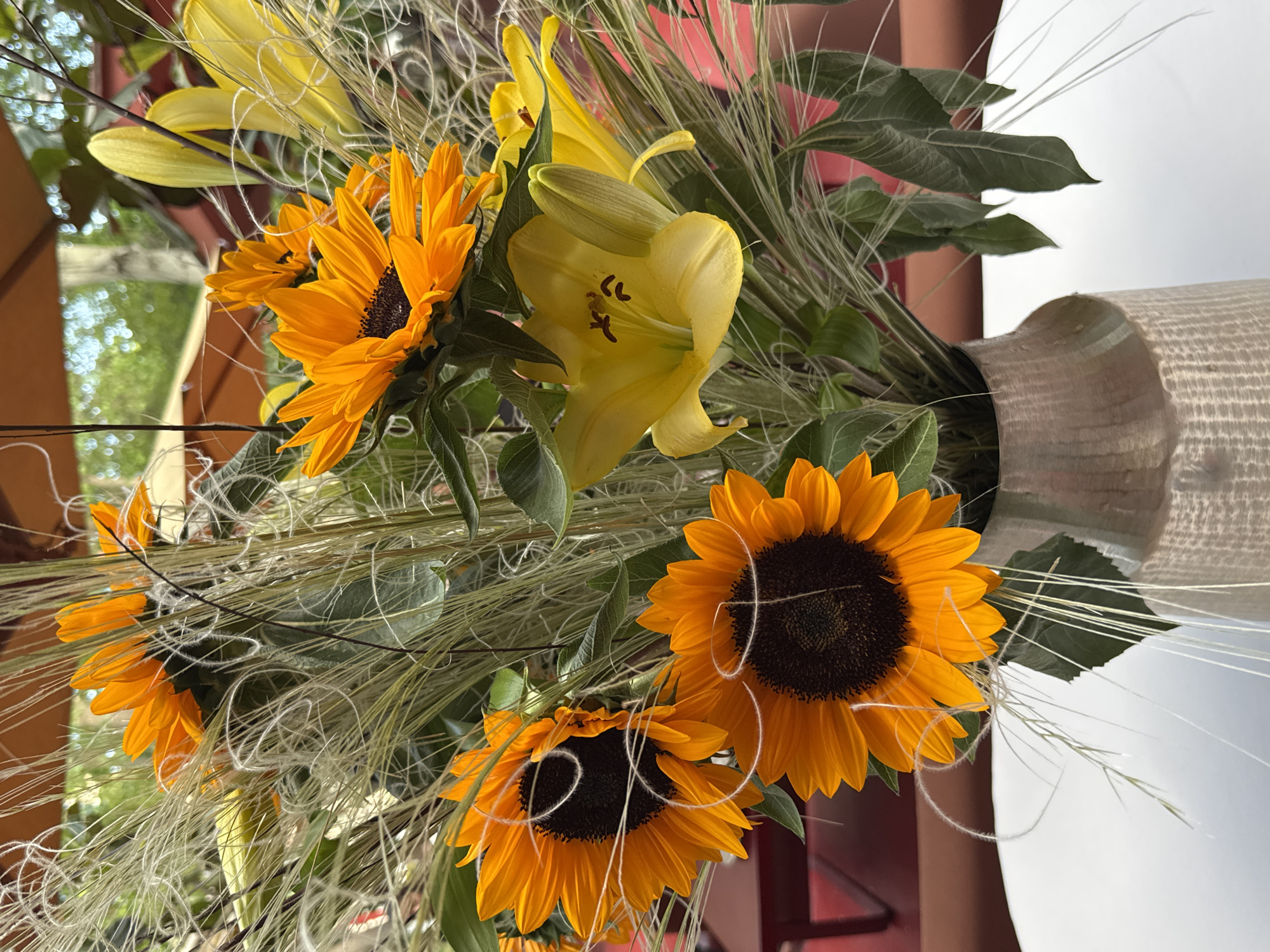 A vase of flowers containing sunflowers and yellow lilies sits on a surface. The vase is textured in light beige or gray. The flowers are arranged with green leafy foliage and decorative wispy strands. The sunflowers are bright orange with dark brown centers. In the background, glimpses of a city scene are visible, suggesting an outdoor setting, possibly in Yerevan, Armenia. A red surface is also visible in the background.