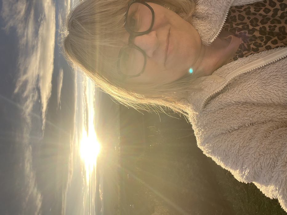 A sunlit selfie features a blonde woman with glasses, wearing a fuzzy beige jacket over a leopard-print top. The bright sun is behind her, creating lens flare. The background includes a sky with clouds and a landscape view. The angle is slightly tilted, with the sun on the lower left side of the frame.