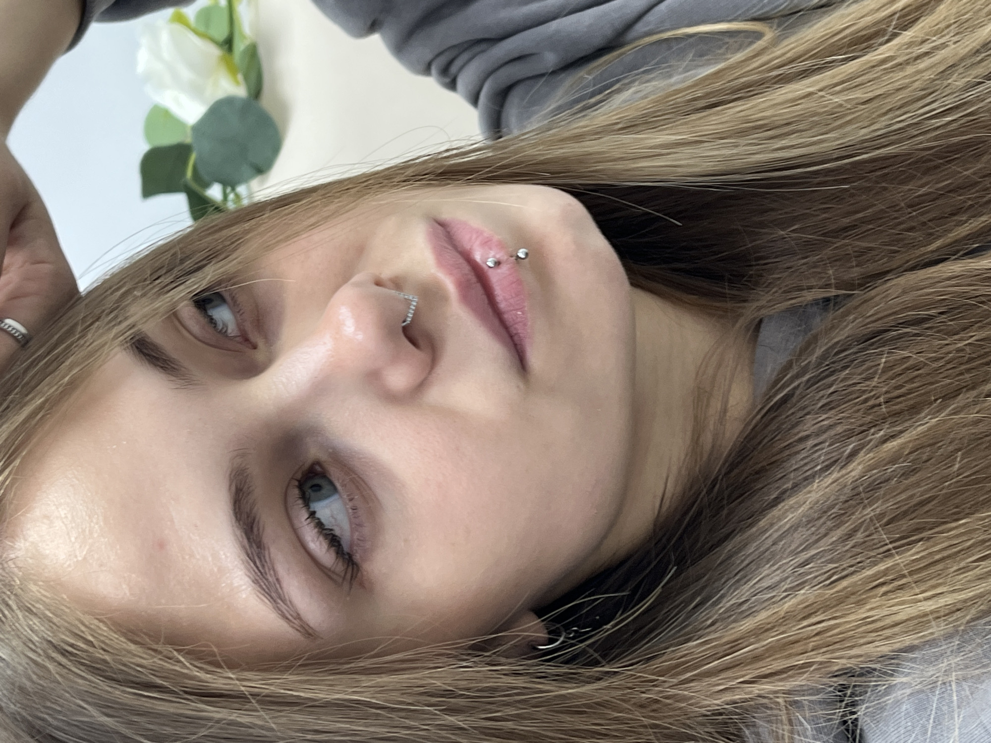 A young woman with light blonde hair is lying down, looking up. She has light blue eyes and fair skin. Her nose is pierced with a small ring and she has a double lip piercing on the right side of her upper lip. She's wearing a gray sweatshirt. A white flower with green leaves is visible in the background. The location in the city of Фоки, Russia cannot be determined from the given information.
