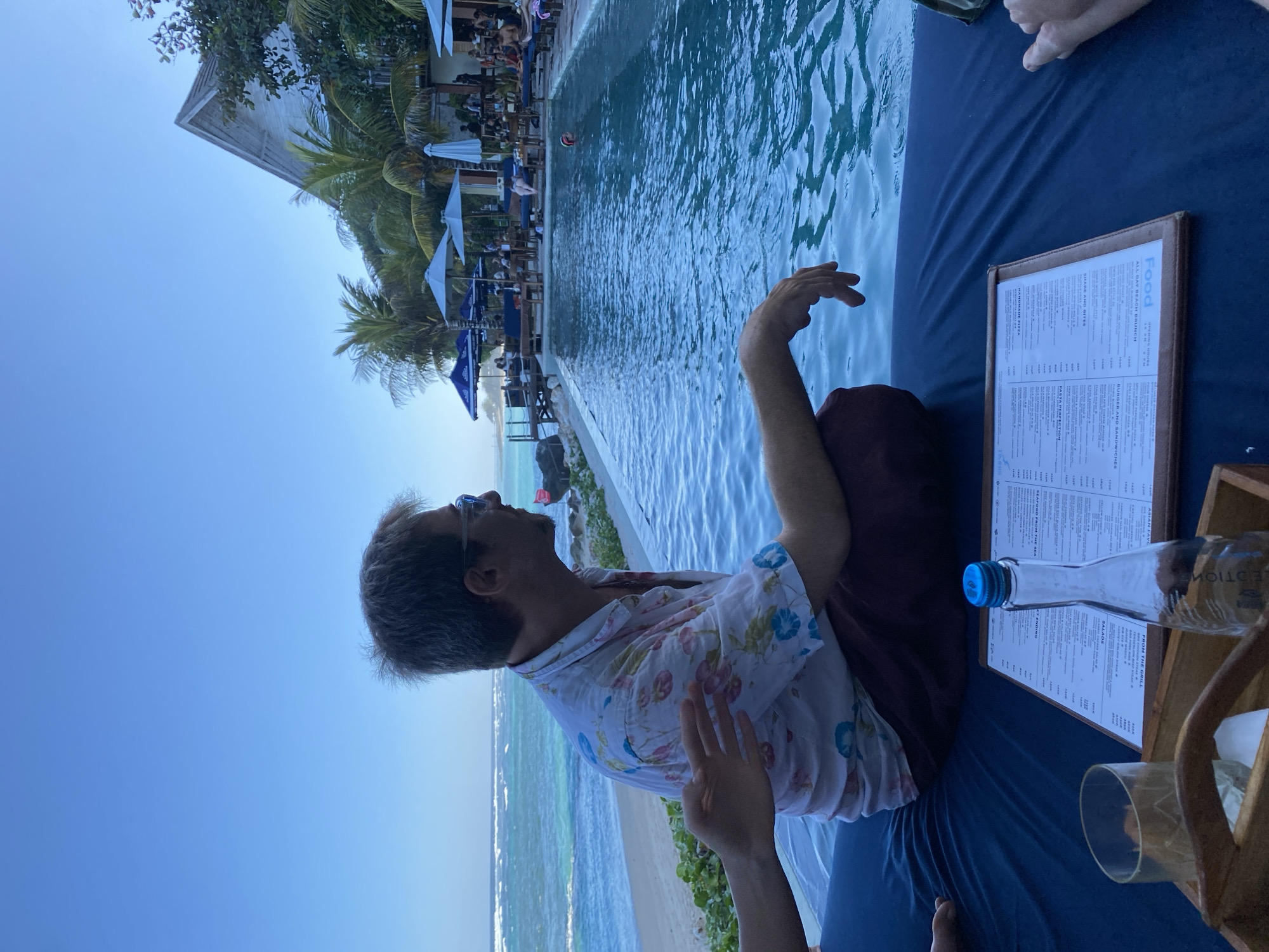In Kuta Selatan, Indonesia, a man with graying hair, wearing glasses and a floral shirt, sits facing away from the camera, looking towards the ocean and a swimming pool. His left arm rests on his lap, while his right hand is extended over the edge of the pool. In the foreground, a menu, a glass with ice, and a water bottle are placed on a dark blue surface. The background features a beach, the ocean with waves, palm trees, umbrellas, and a building. Several people are visible in the background, some near the pool and others in the seating area. The sky is clear and blue.