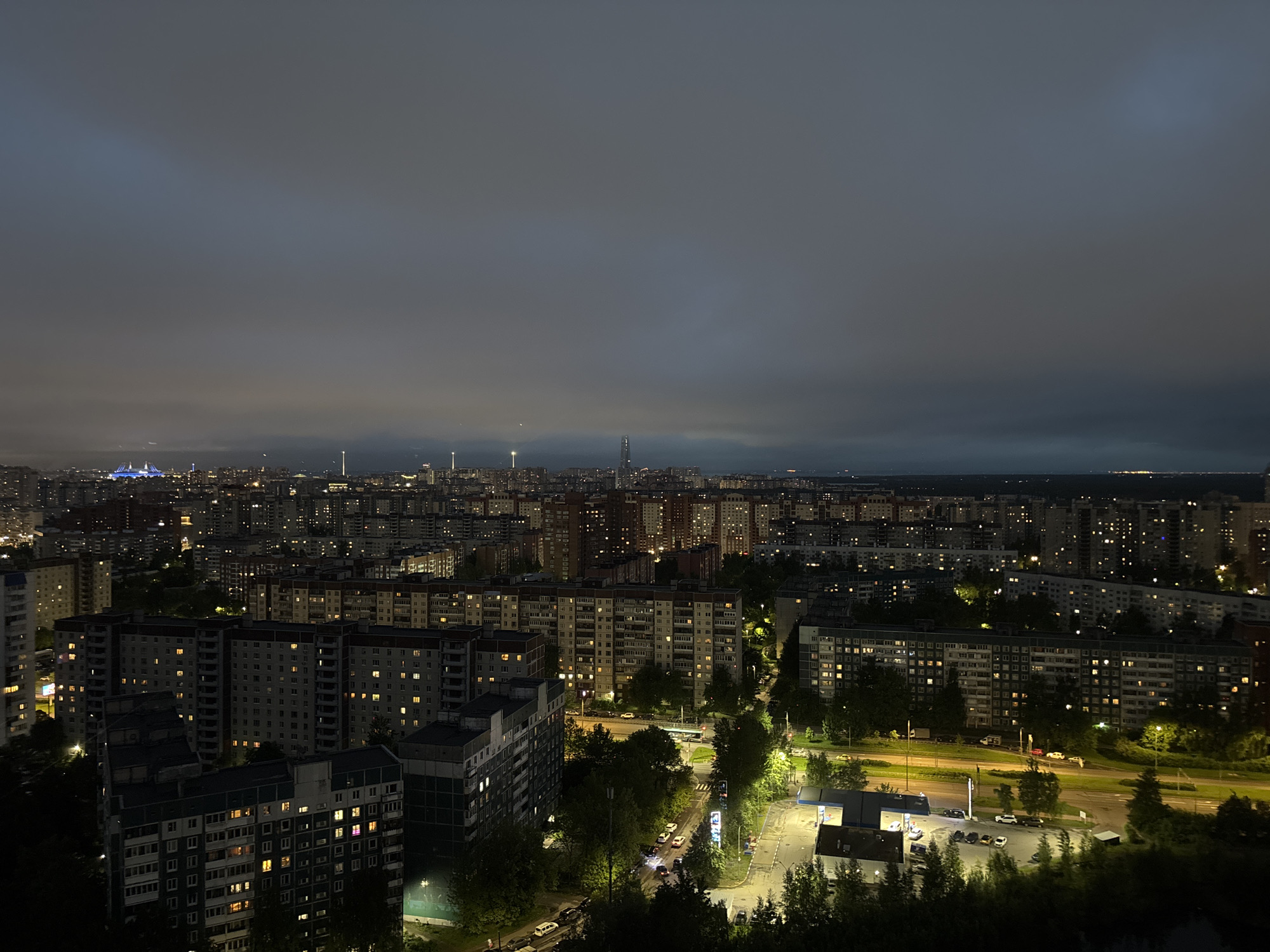 The image captures a nighttime view of an urban area. Numerous buildings, likely residential apartment blocks, are visible in the foreground and middle ground, illuminated by scattered lights. A road runs through the scene, with cars and streetlights visible. In the background, a large city skyline stretches under a cloudy night sky, with several prominent structures including a tall spire and a stadium with blue lights. The scene appears to be in a city, but the exact location within the city is unknown.