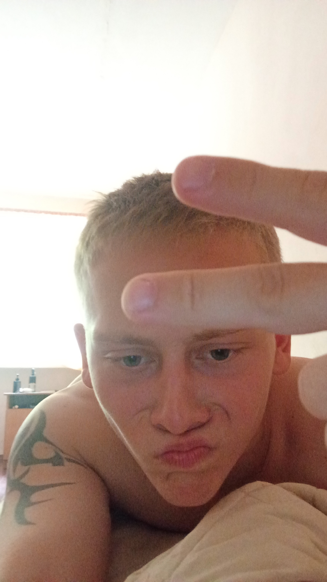 A young male, possibly a teenager, is lying down and taking a selfie. He has light brown hair and blue eyes. His mouth is pursed, and he is holding two fingers up to his forehead in a peace sign gesture. He is wearing a tattoo on his left shoulder. The background is out of focus, but appears to be a bedroom with a dresser and some bottles on it. The lighting suggests it is daytime. The location is unknown, and cannot be determined to be Tomsk, Russia.