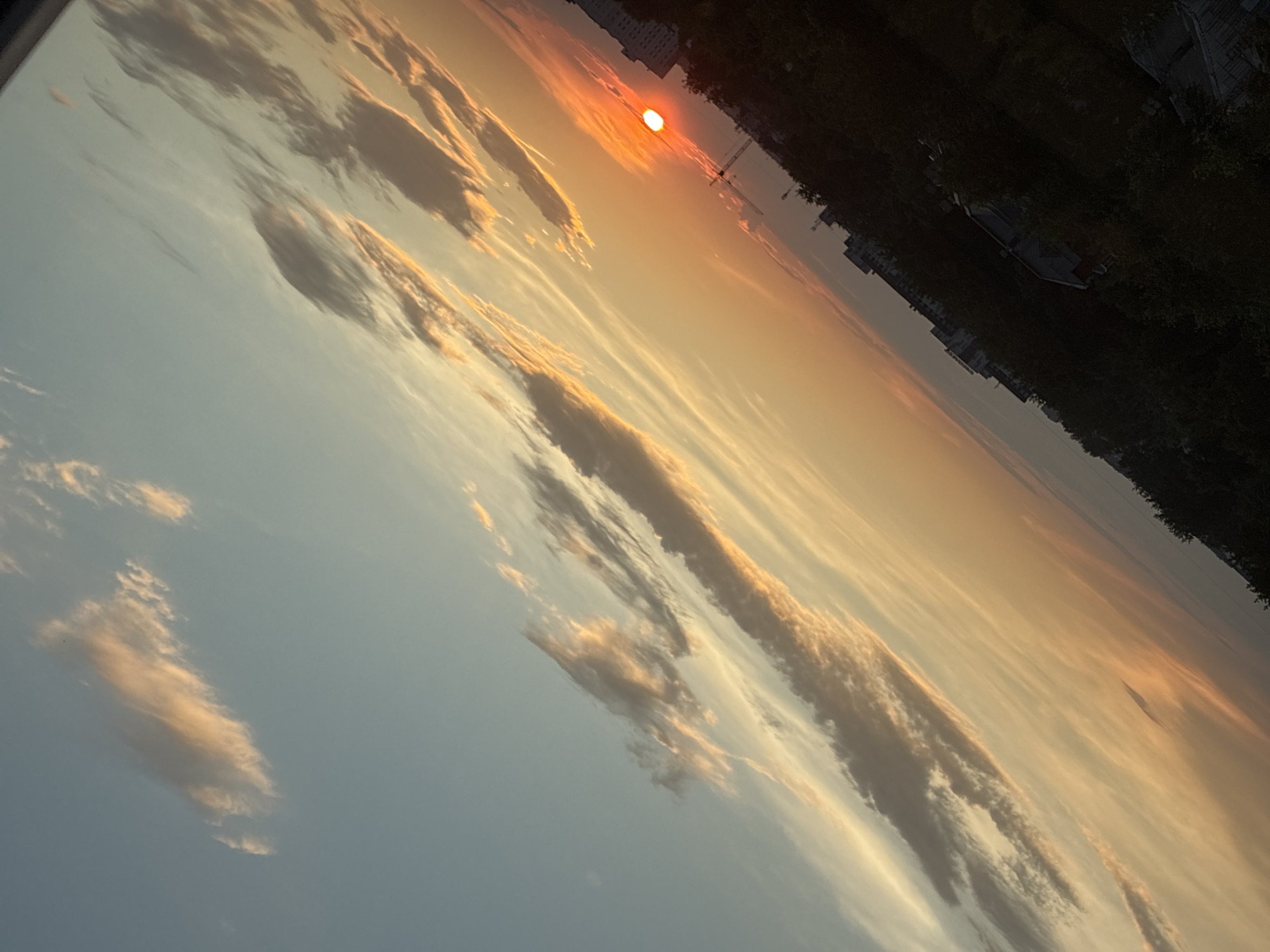 This is a diagonal, outdoor, medium-shot photograph of a sunset over an urban environment. The sun, a bright orange disc, is low on the horizon, casting a warm glow across the sky. Wispy clouds are scattered across the sky, painted with hues of orange, pink, and yellow. In the foreground, silhouetted against the sunset, are buildings and trees, suggesting a city landscape. The image is tilted, giving a dynamic perspective to the scene. The location is Unknown, Unknown.