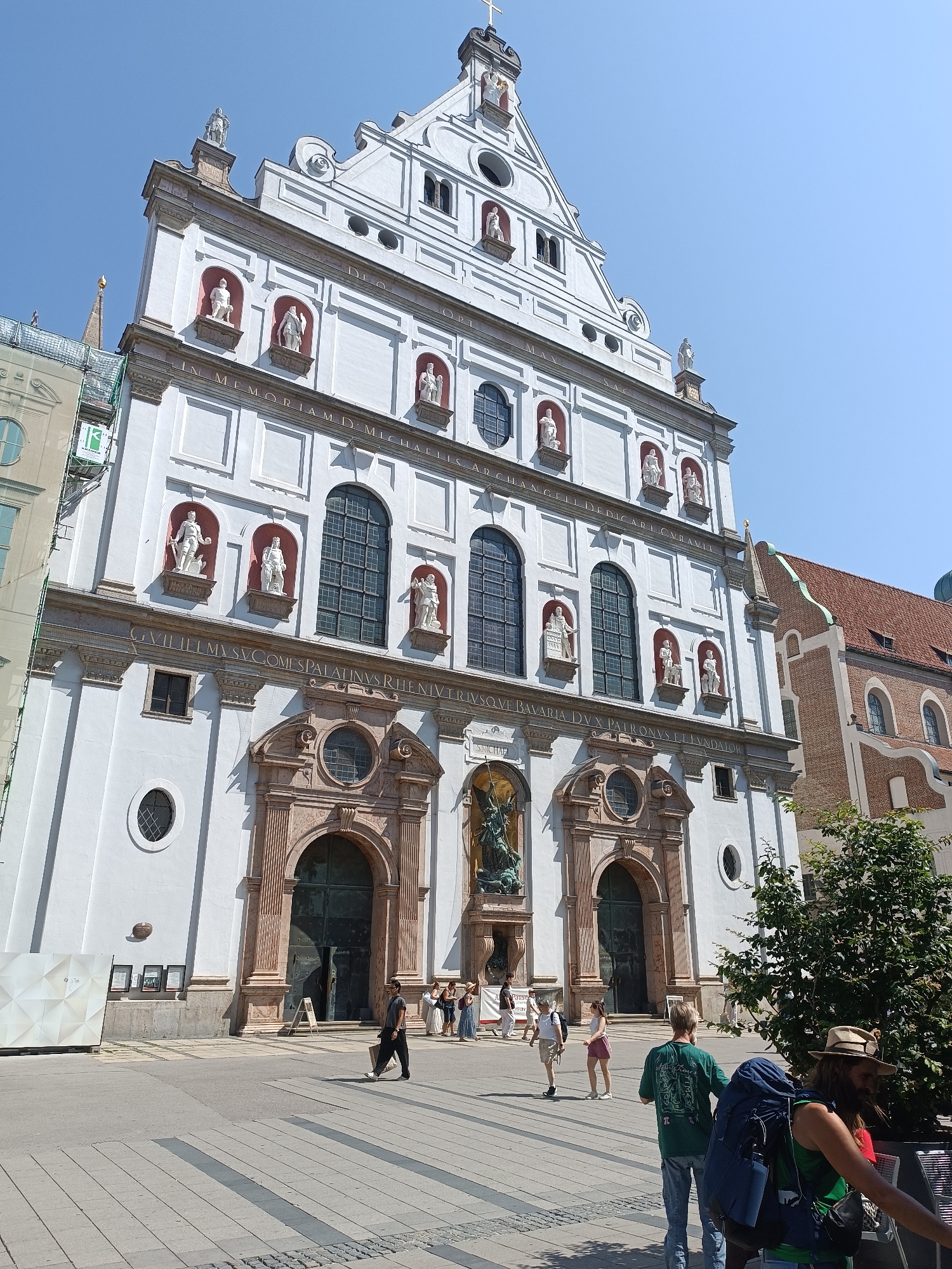 The image shows the St. Michael's Church in Munich, Germany, on a clear day. The church is a large white Renaissance building with a stepped gable, decorated with statues in niches. In front of the church, a paved plaza is visible with several people walking. Some of the people are carrying backpacks, suggesting they might be tourists. A large green tree is located on the right side of the image, and a smaller building with a red tiled roof is visible behind the church. The scene is brightly lit by the sun, casting shadows on the plaza.