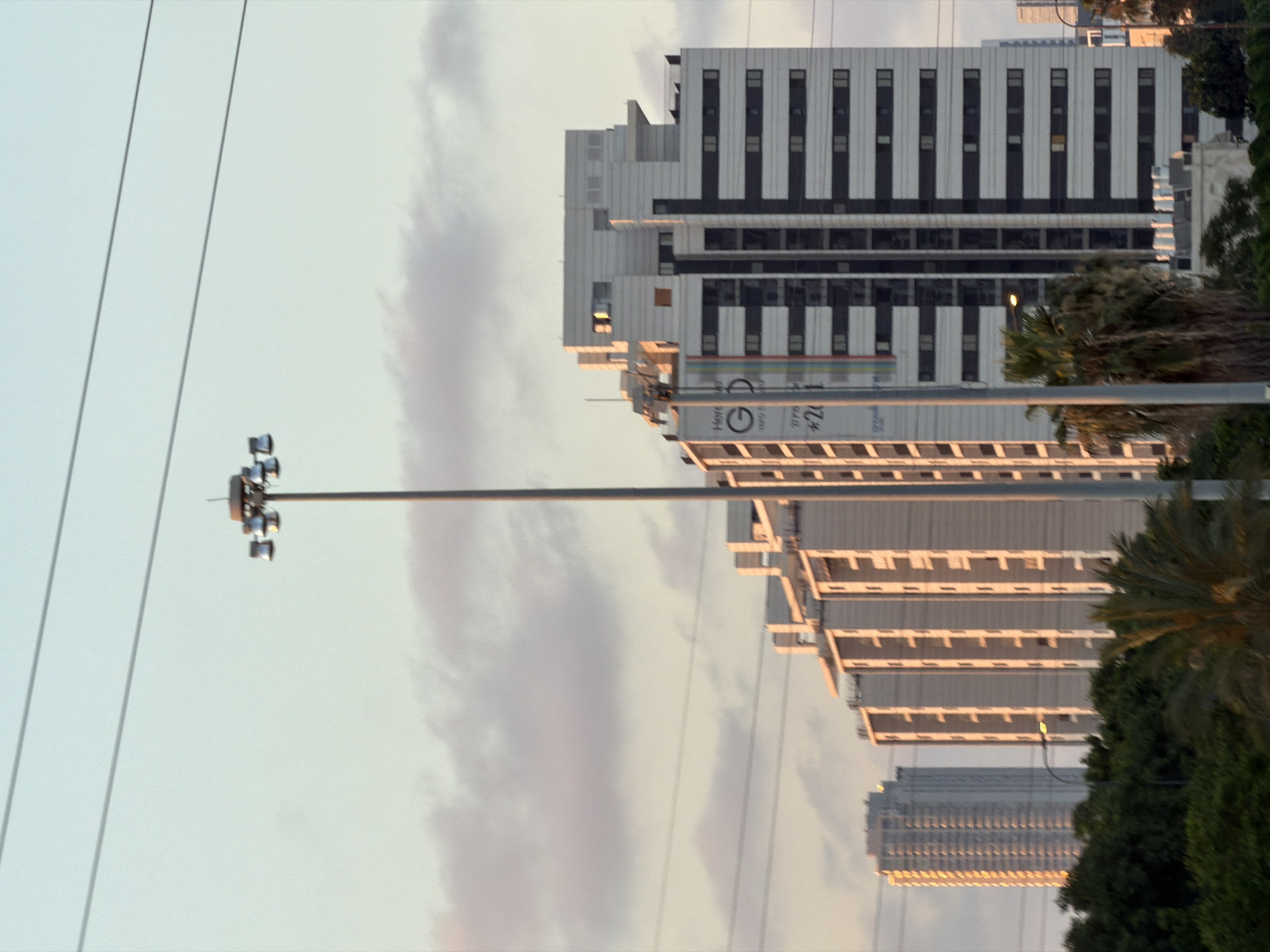 The image captures an urban skyline in Tel Aviv, Israel, at what appears to be either dawn or dusk, given the warm lighting on the buildings and the soft, diffused light in the sky. Dominating the foreground is a tall, modern building with a distinctive facade featuring alternating dark and white horizontal bands, creating a rhythmic visual pattern. The building has multiple floors, each with a series of windows. To the left of this central structure, another taller, slender building is visible, also illuminated by the low-angle sun, showcasing rows of balconies or recessed windows.
A large, multi-headed light fixture on a tall pole stands prominently in the middle ground, suggesting it's a streetlamp or part of a stadium lighting system. In the lower portion of the image, lush green trees and palm trees form a verdant contrast to the concrete and glass structures.
The sky is a soft gradient of pale blue and pinkish hues, with wispy clouds scattered across it. Several power lines are visible, stretching horizontally across the frame, indicating the presence of electrical infrastructure. A large banner or sign is attached to the central building, partially obscured, with visible text including "GD" and what appears to be a phone number. The overall impression is one of a modern city with significant development, captured during a tranquil moment of the day.
