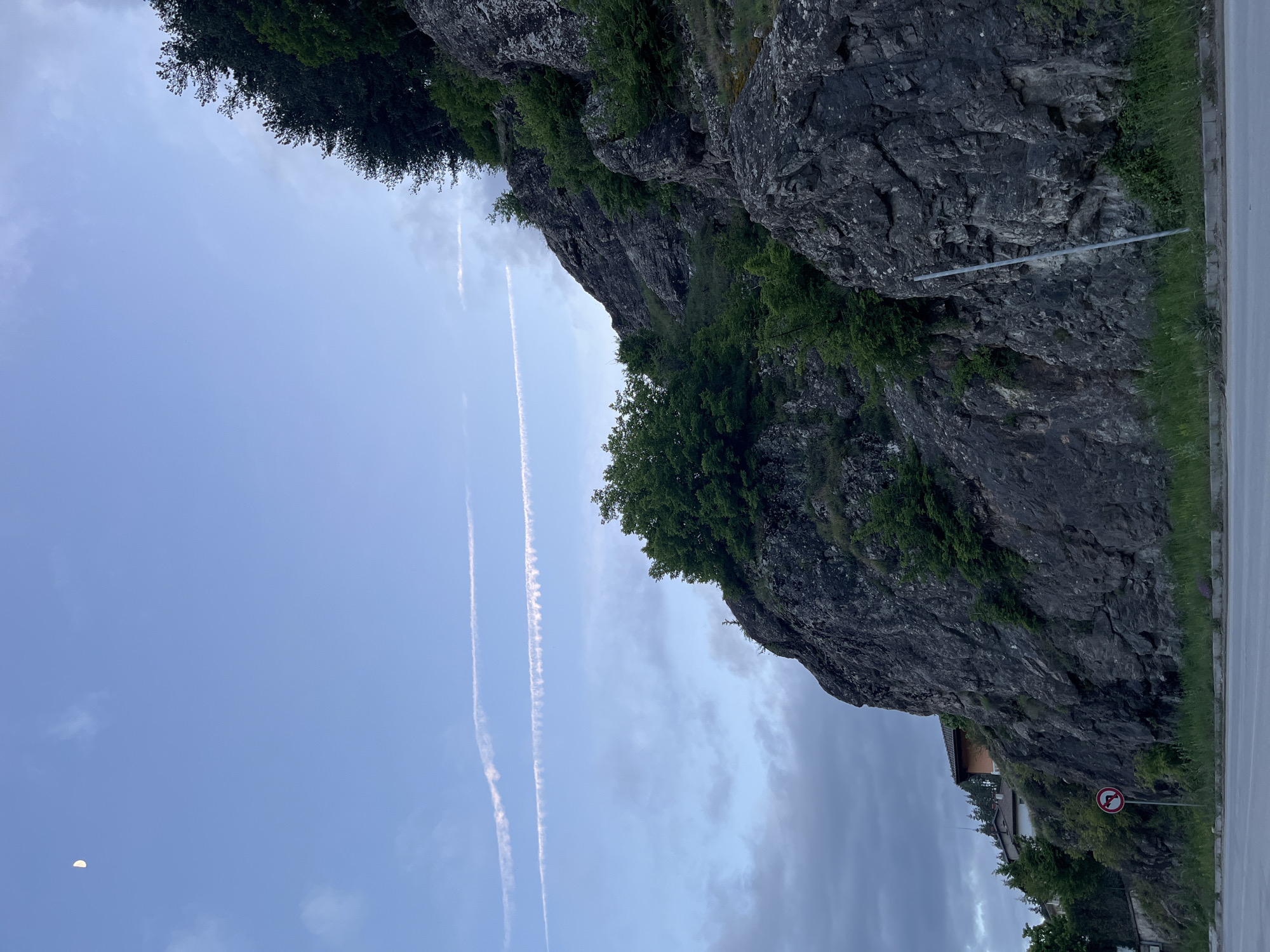 The image shows a vertical view of a rocky cliff with vegetation on its surface, likely located alongside a road in Rudozem, Bulgaria. The sky is partially cloudy, with two prominent contrails from aircraft. At the lower right edge of the frame is a road with a "no pedestrians" sign. There is a hint of buildings/housing nestled near the base of the cliff. A crescent moon is visible in the lower left portion of the sky.