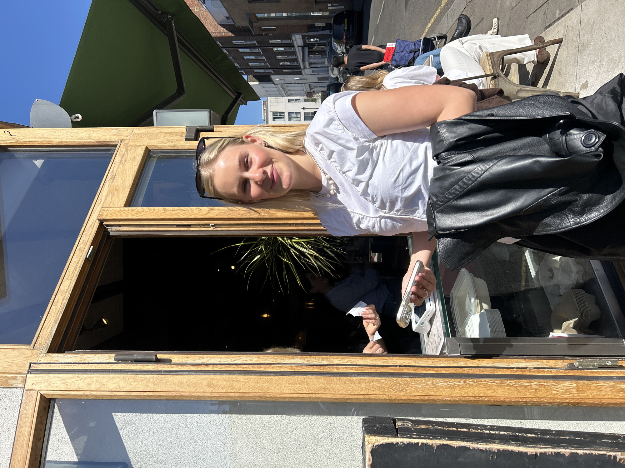 A fair-skinned young woman stands outside a building in the city of London, United Kingdom. She wears a white blouse with frills and a pearl necklace. Sunglasses are perched on her head. A black leather jacket is draped over her shoulder. She is looking at the camera with a slight smile.
The woman is standing next to an open window of a building. Inside the window, a person is visible holding a receipt and a phone. There are also takeout containers and napkins.
Outside, other people are seated at tables on the sidewalk. Buildings line the street. An awning shades the window.