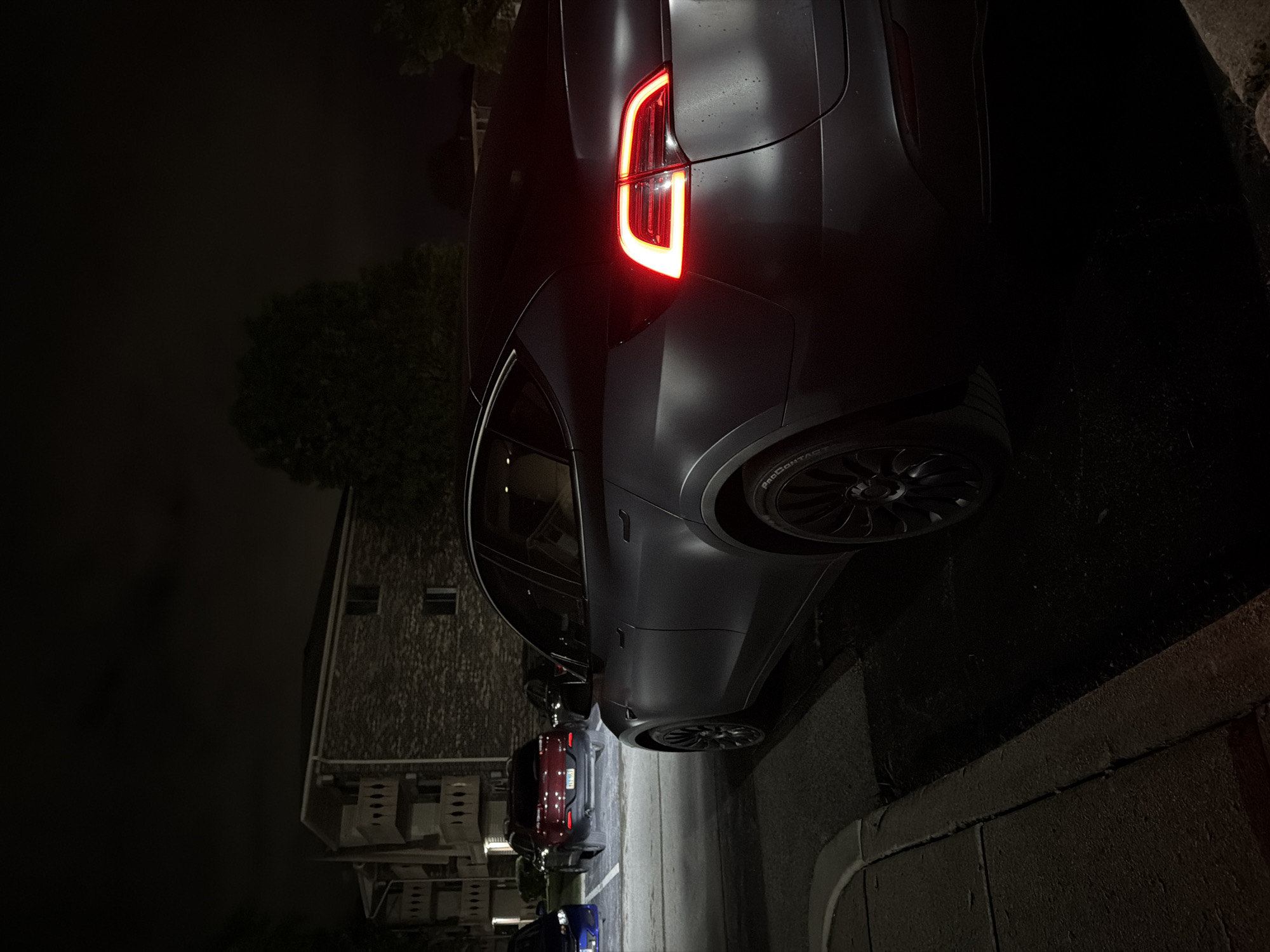 A matte gray Tesla Model Y is parked at night in a residential area in Vernon Hills, Illinois. The car's rear taillights are illuminated, casting a red glow. The vehicle is parked in a lot next to a curb and asphalt pavement. In the background, an apartment building with balconies is visible, along with other parked cars, including a red SUV and a blue sedan. Trees and foliage are also present, partially obscuring the building and sky. The scene is dimly lit, characteristic of nighttime conditions.